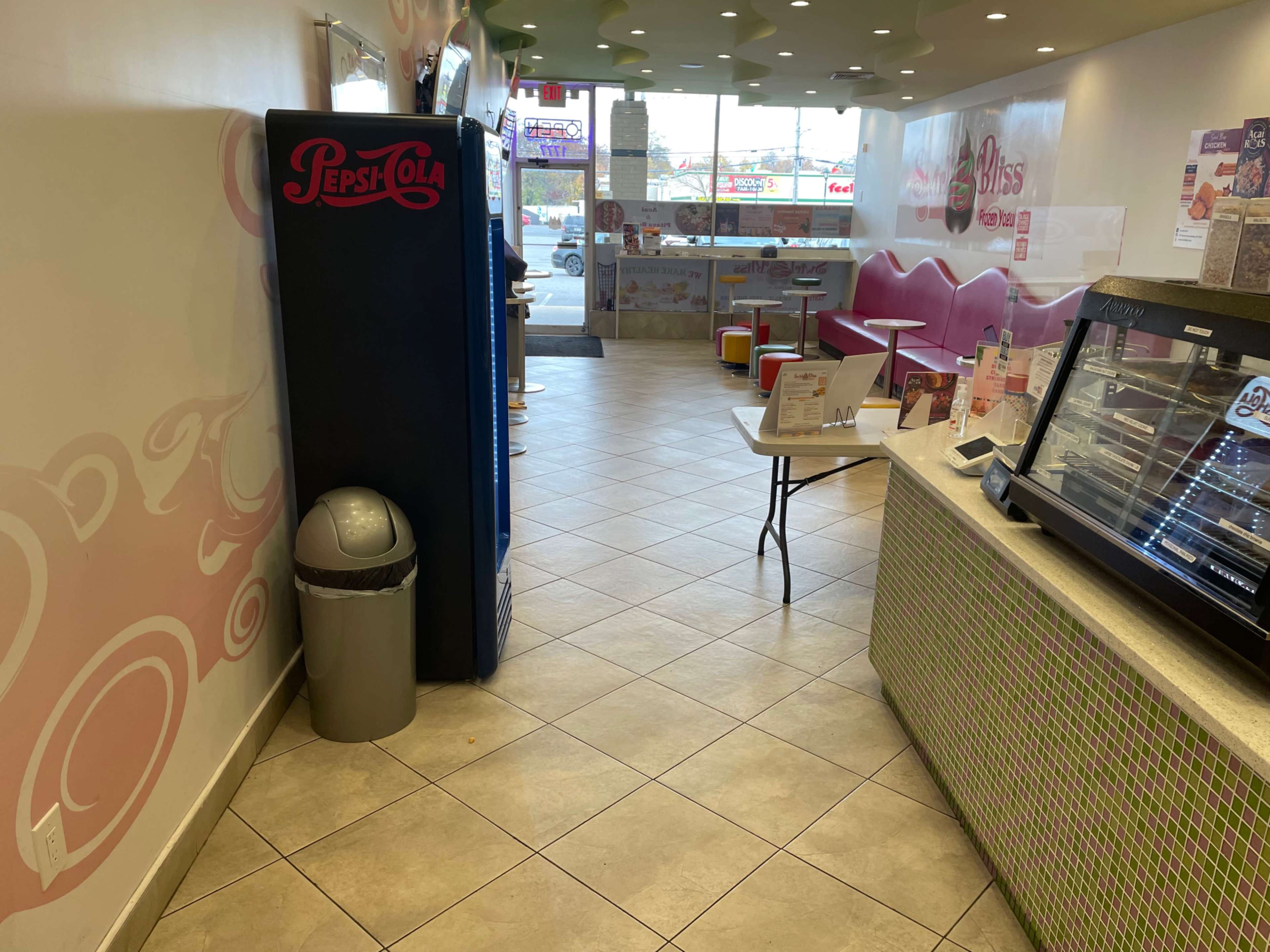 A brightly lit café interior featuring a Pepsi vending machine, a table with chairs, and a display case against a wall.