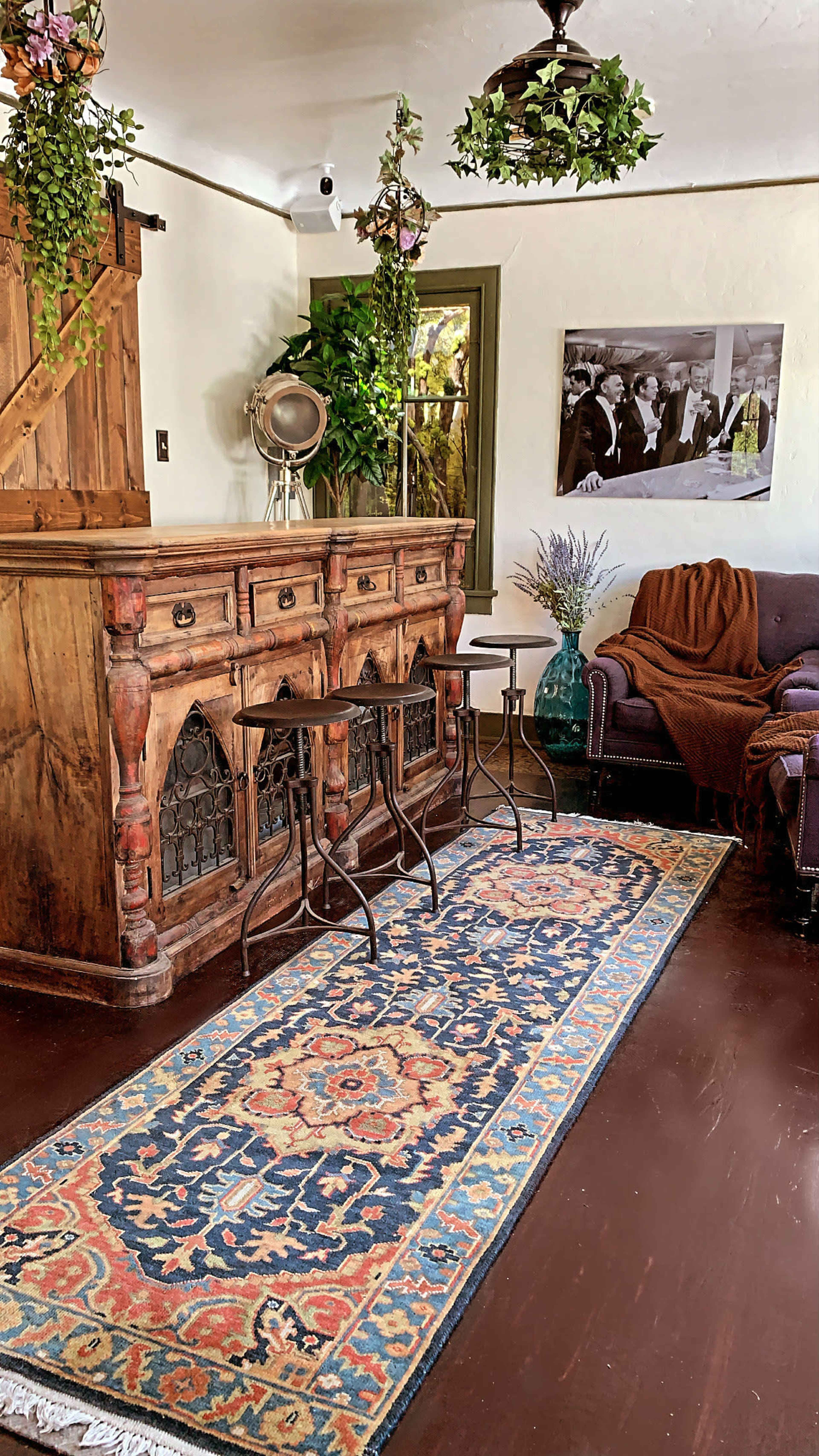 The interior features a rustic wooden sideboard, two metal-legged tables, a patterned area rug, and a cozy seating arrangement with plants and a black-and-white photograph on the wall.