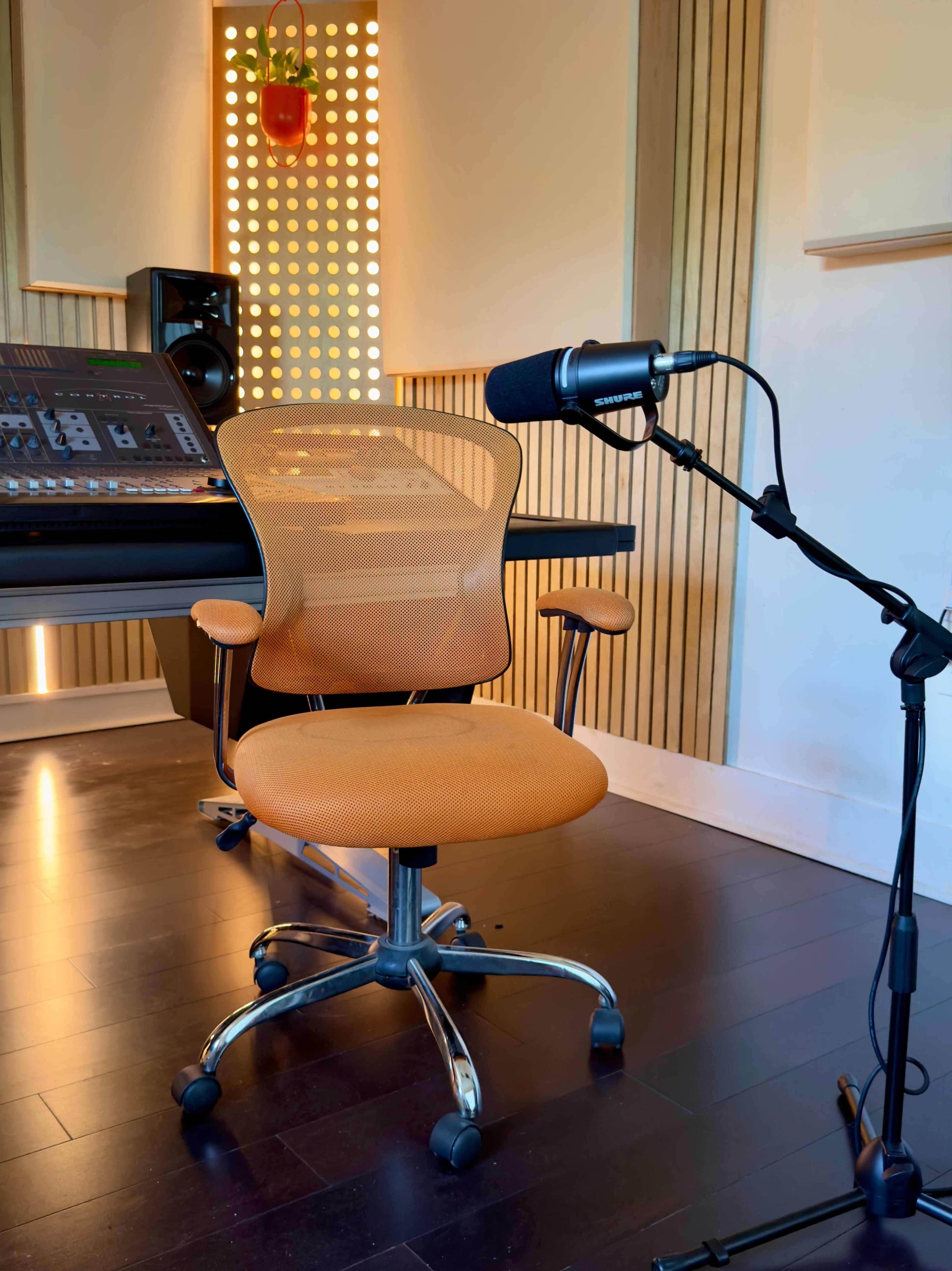 A vibrant orange office chair is positioned next to a microphone on a stand, with sound equipment and wooden paneling in the background of a recording studio.