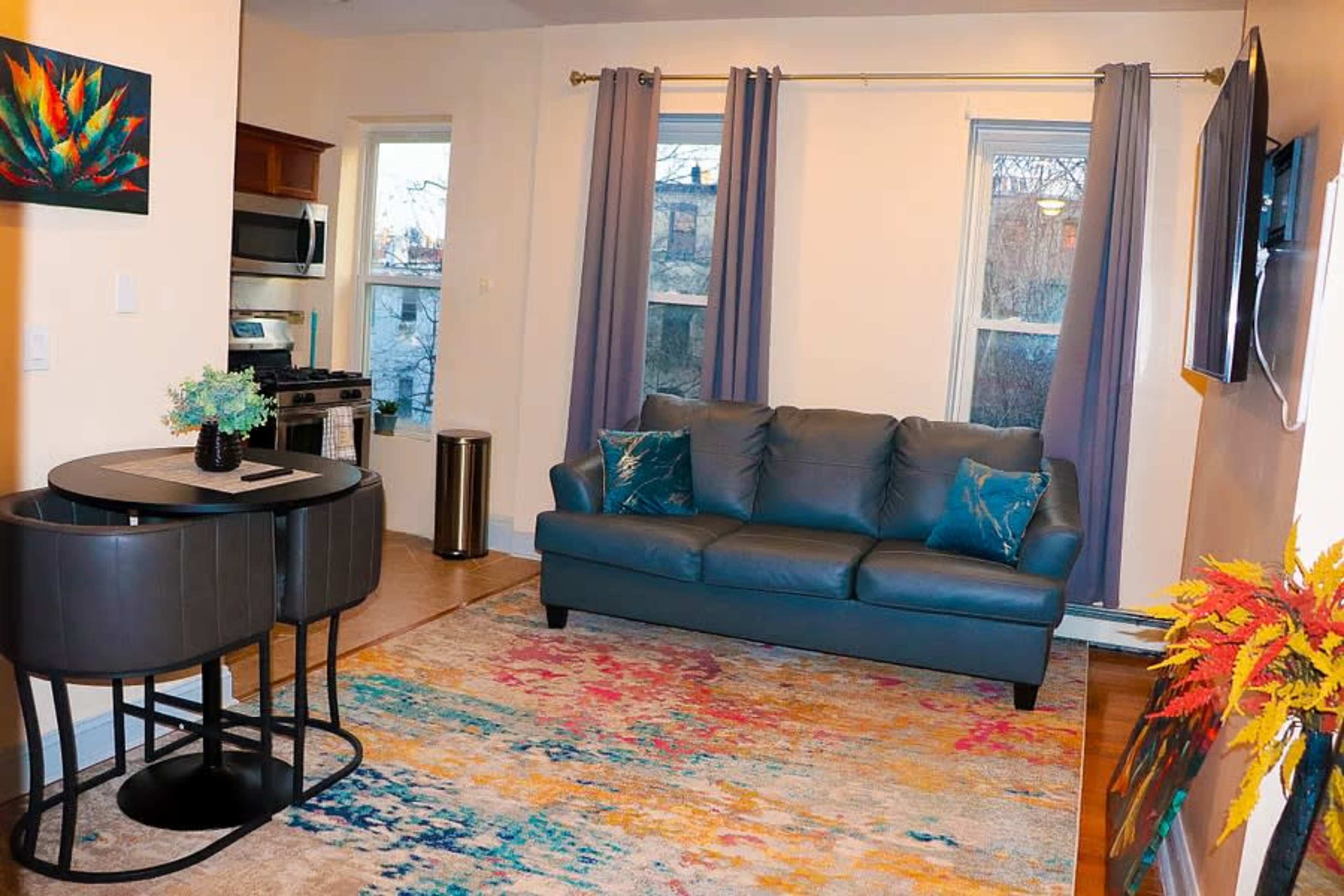 The image shows a modern living room featuring a blue sofa, a small dining table, and colorful decor on the floor and walls.