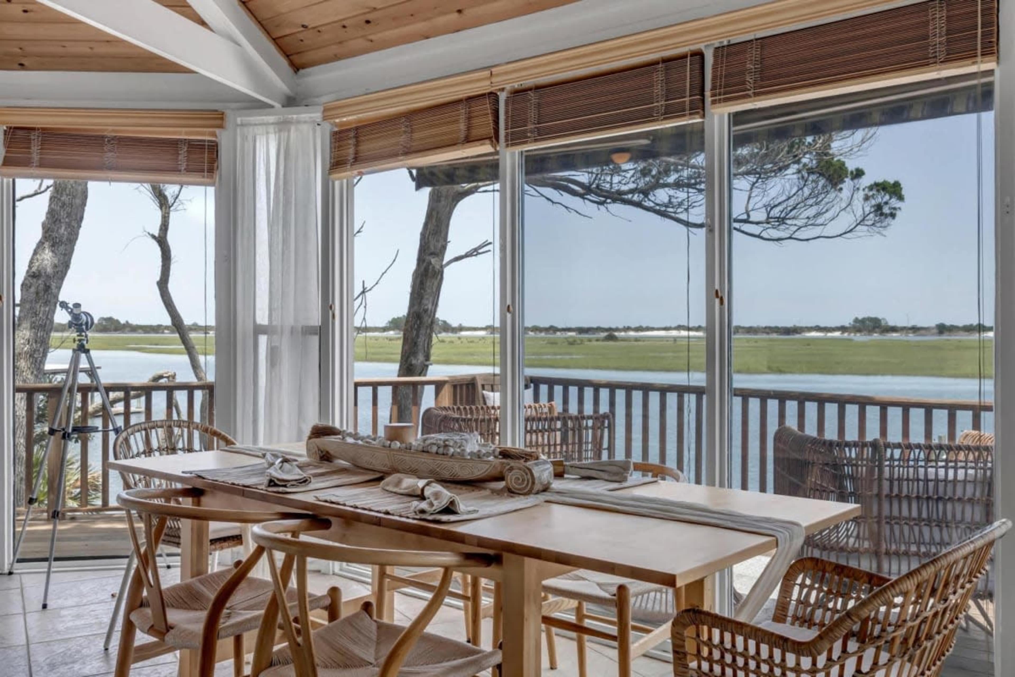 A dining area features a wooden table set with linens and overlooks a peaceful view of a river through large glass windows, framed by wooden beams and wicker furniture.