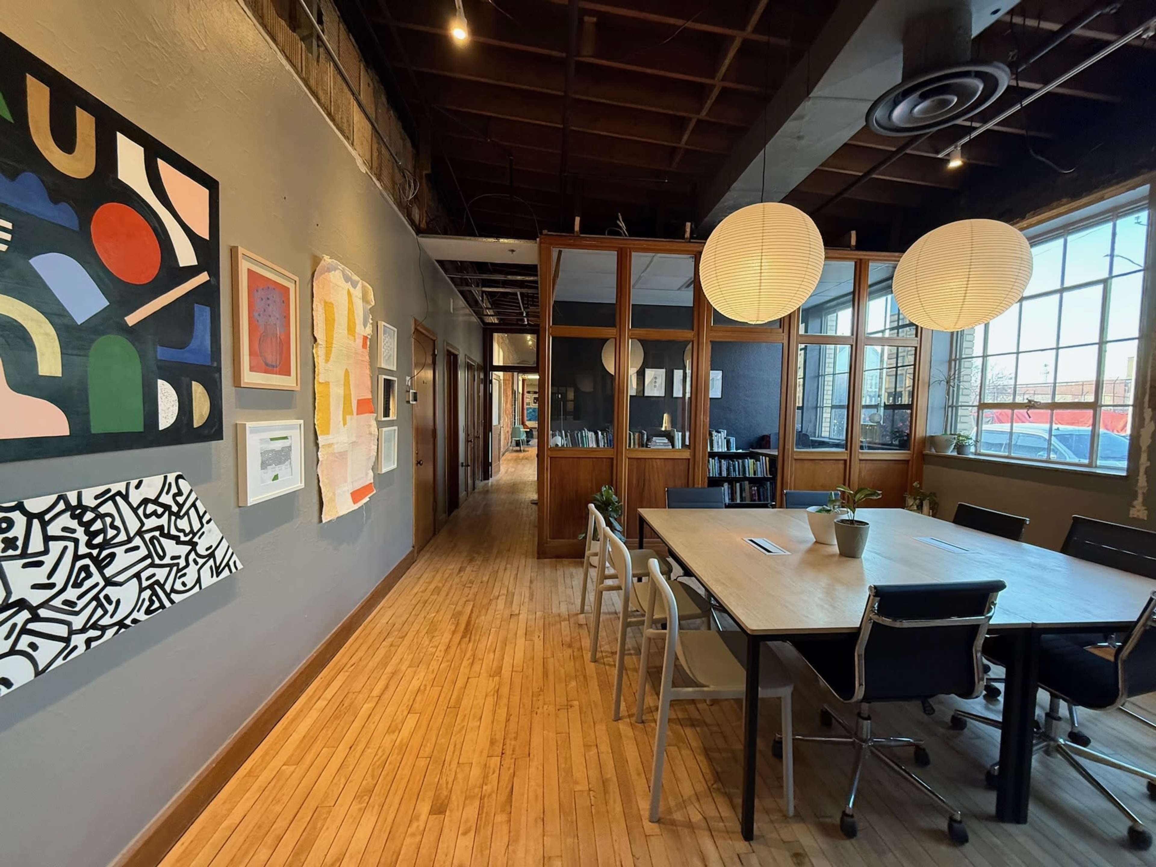 The image shows a brightly lit, modern office space featuring a long table surrounded by chairs, with a gallery wall of colorful artwork on one side and large windows providing natural light.