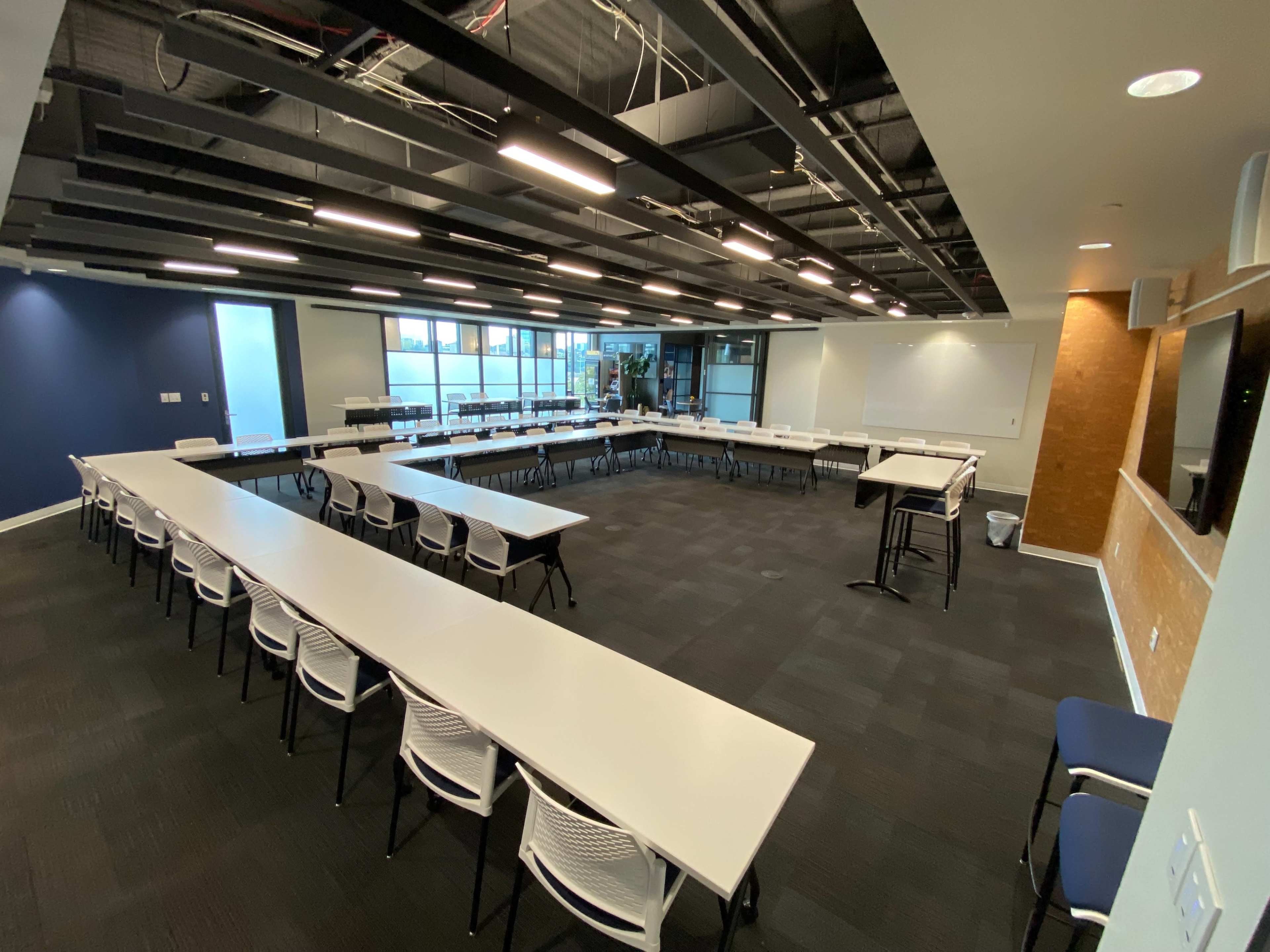 A modern conference room features multiple tables arranged in a U-shape, with white walls and large windows allowing natural light.