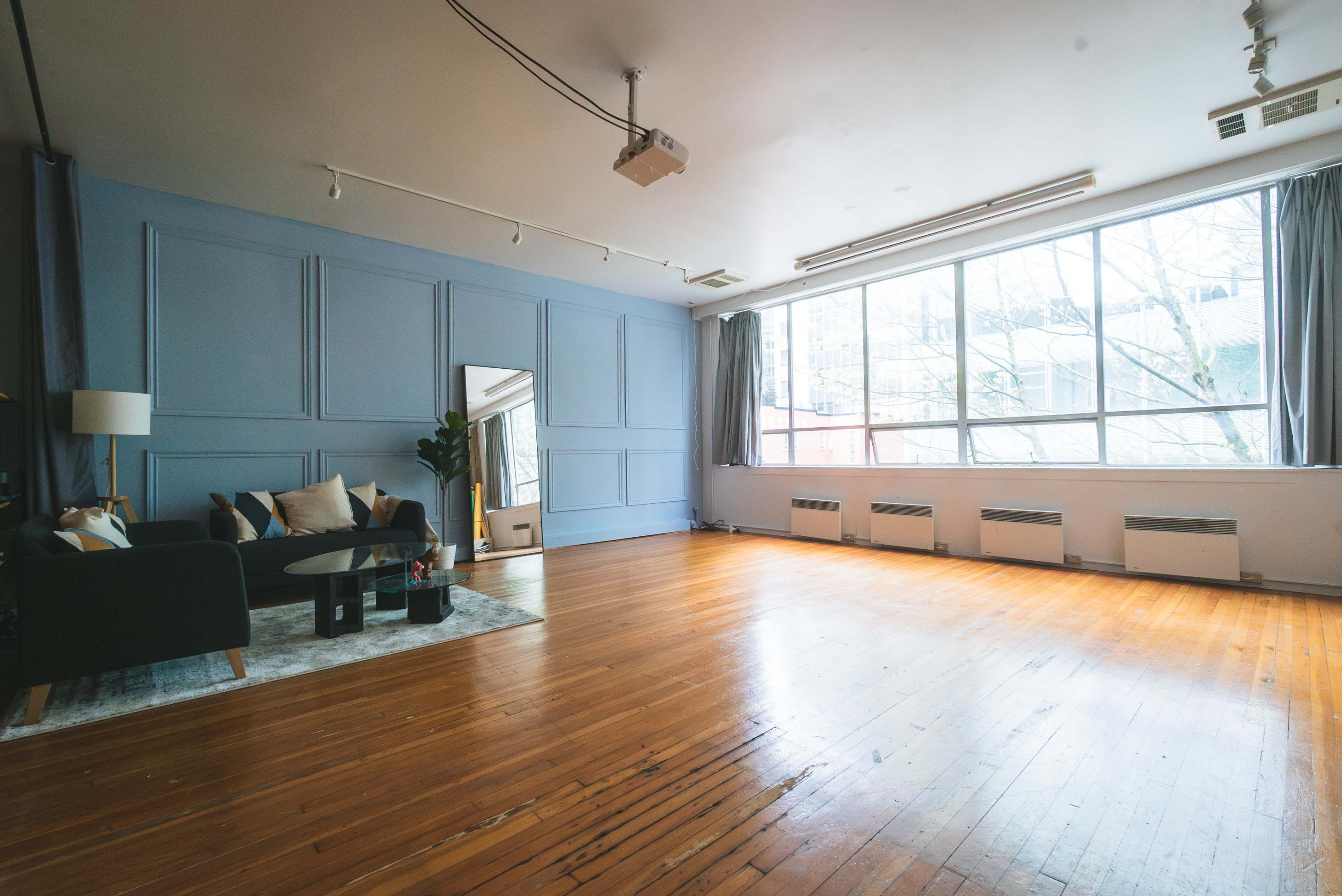 The image shows a spacious room with wooden flooring, large windows, and minimalist furnishings including a sofa and a floor lamp.