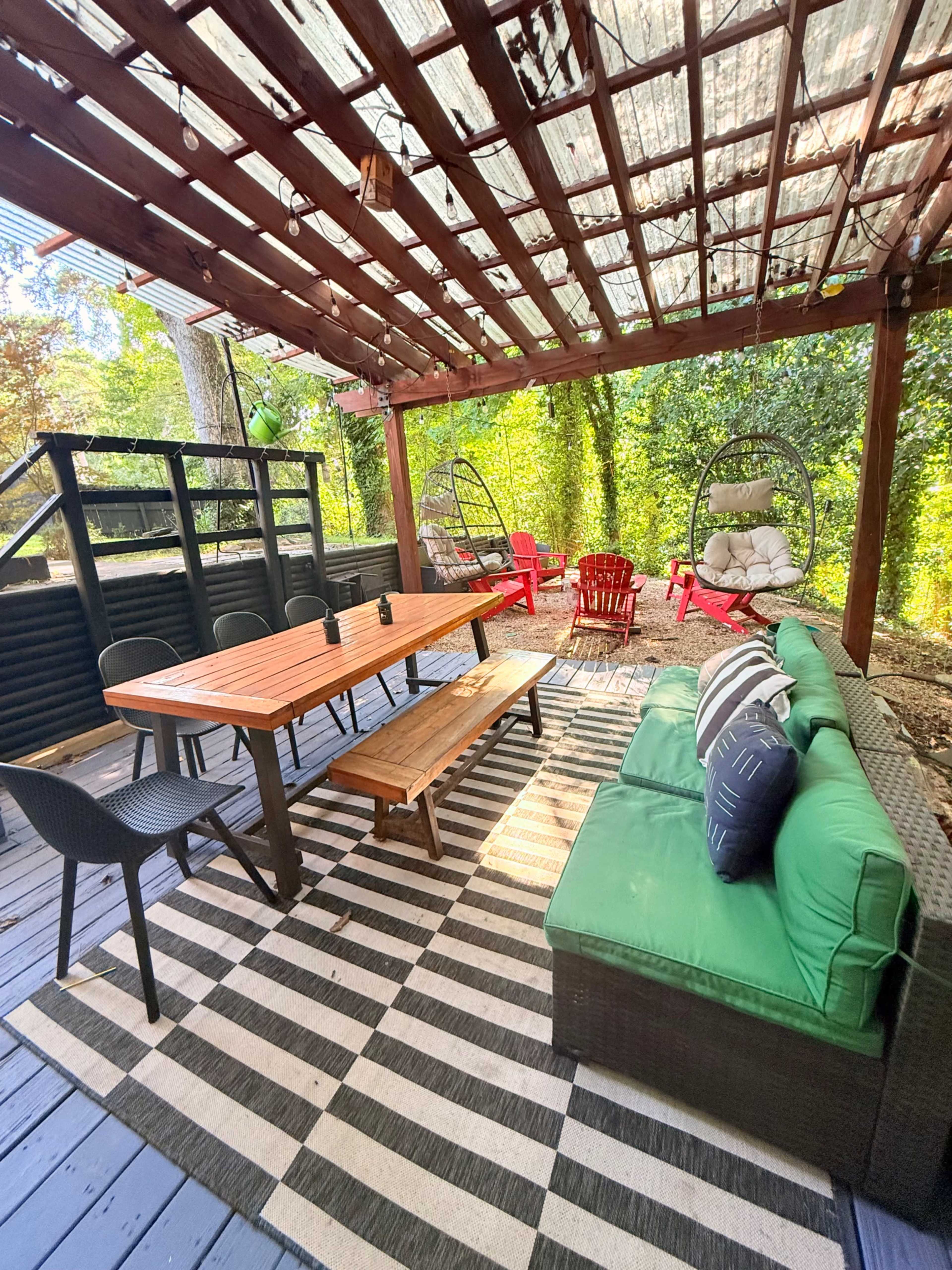 The image shows a shaded outdoor patio featuring a wooden table with benches, a green sectional sofa, and two hanging chairs, all surrounded by greenery.