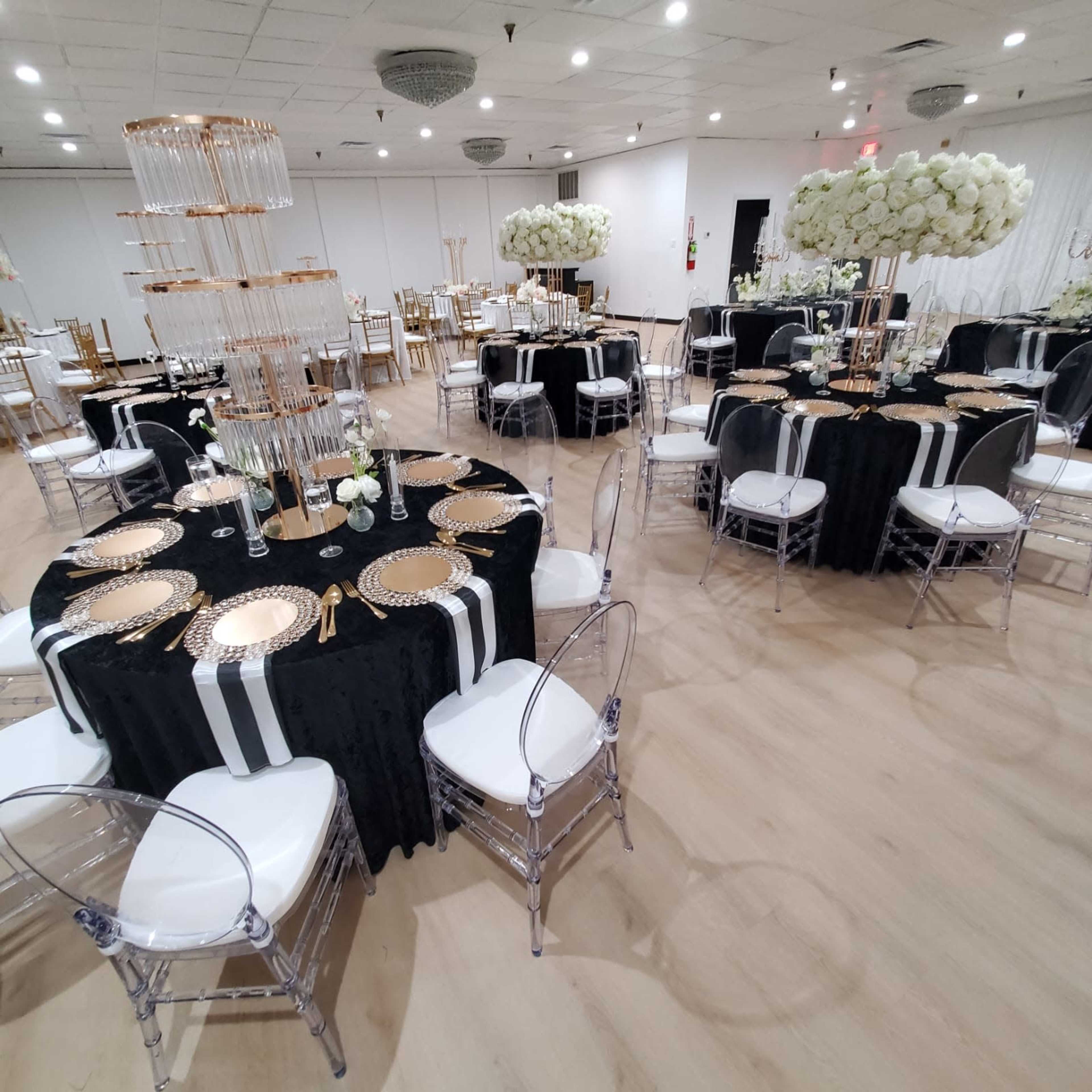 The image shows a banquet hall set for an event with elegantly arranged tables featuring black tablecloths, gold dinnerware, and floral centerpieces.