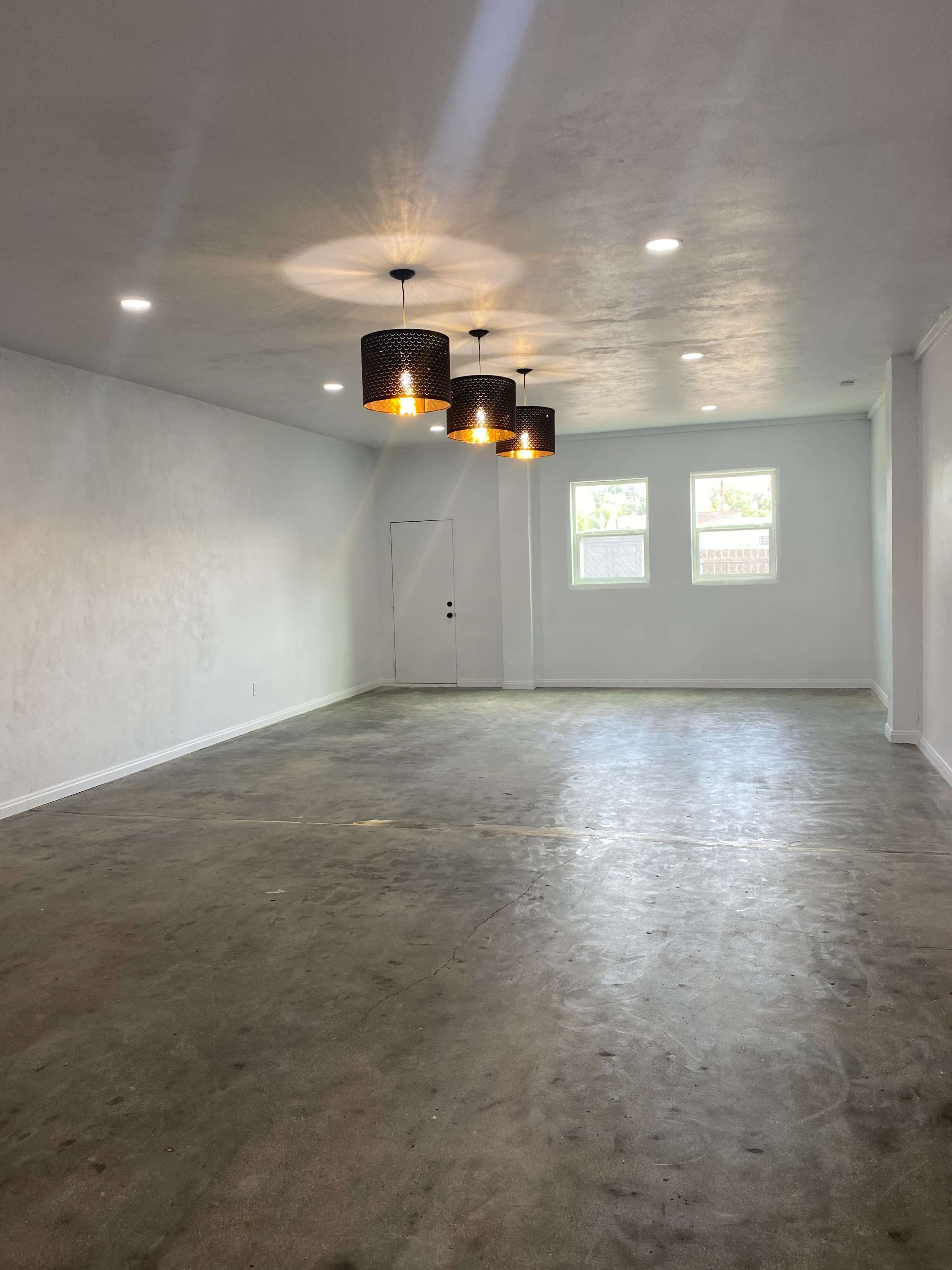 The image shows a spacious, empty room with concrete flooring, white walls, and three pendant lights hanging from the ceiling.