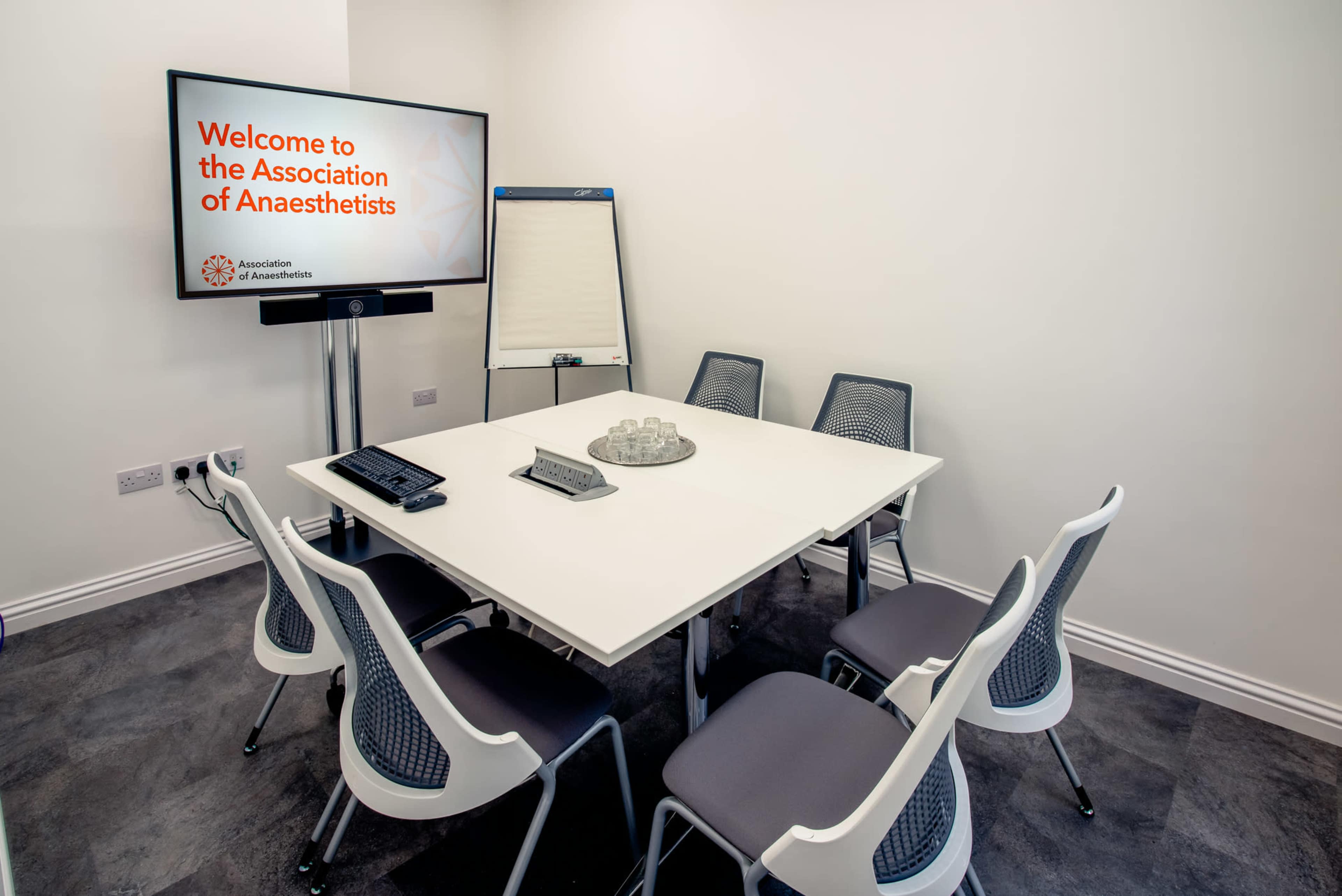 A modern conference room features a large screen displaying a welcome message, a whiteboard, and a rectangular table surrounded by six chairs.