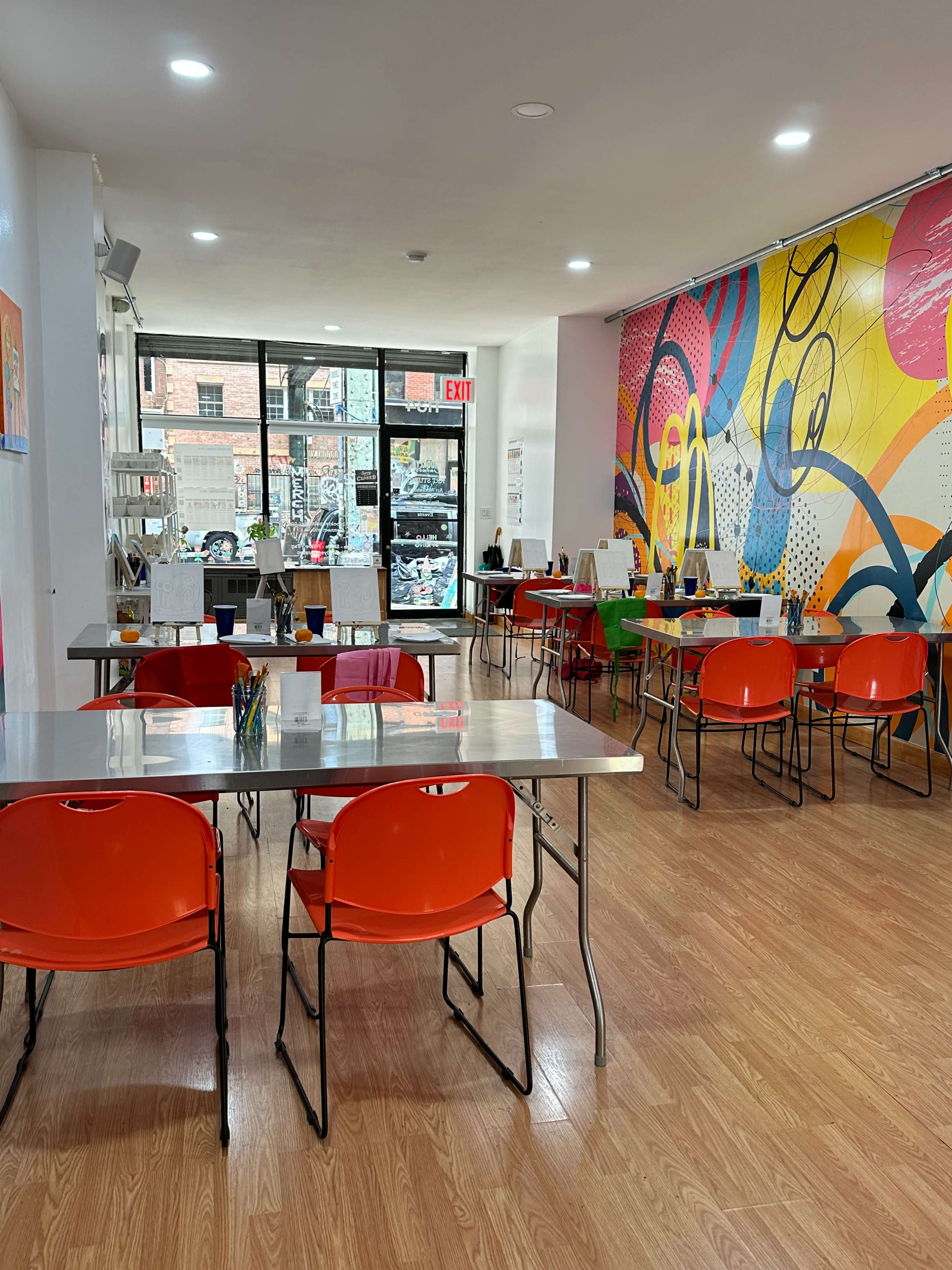 A brightly colored interior of a restaurant with several tables and orange chairs, large windows in the background, and vibrant murals on the walls.