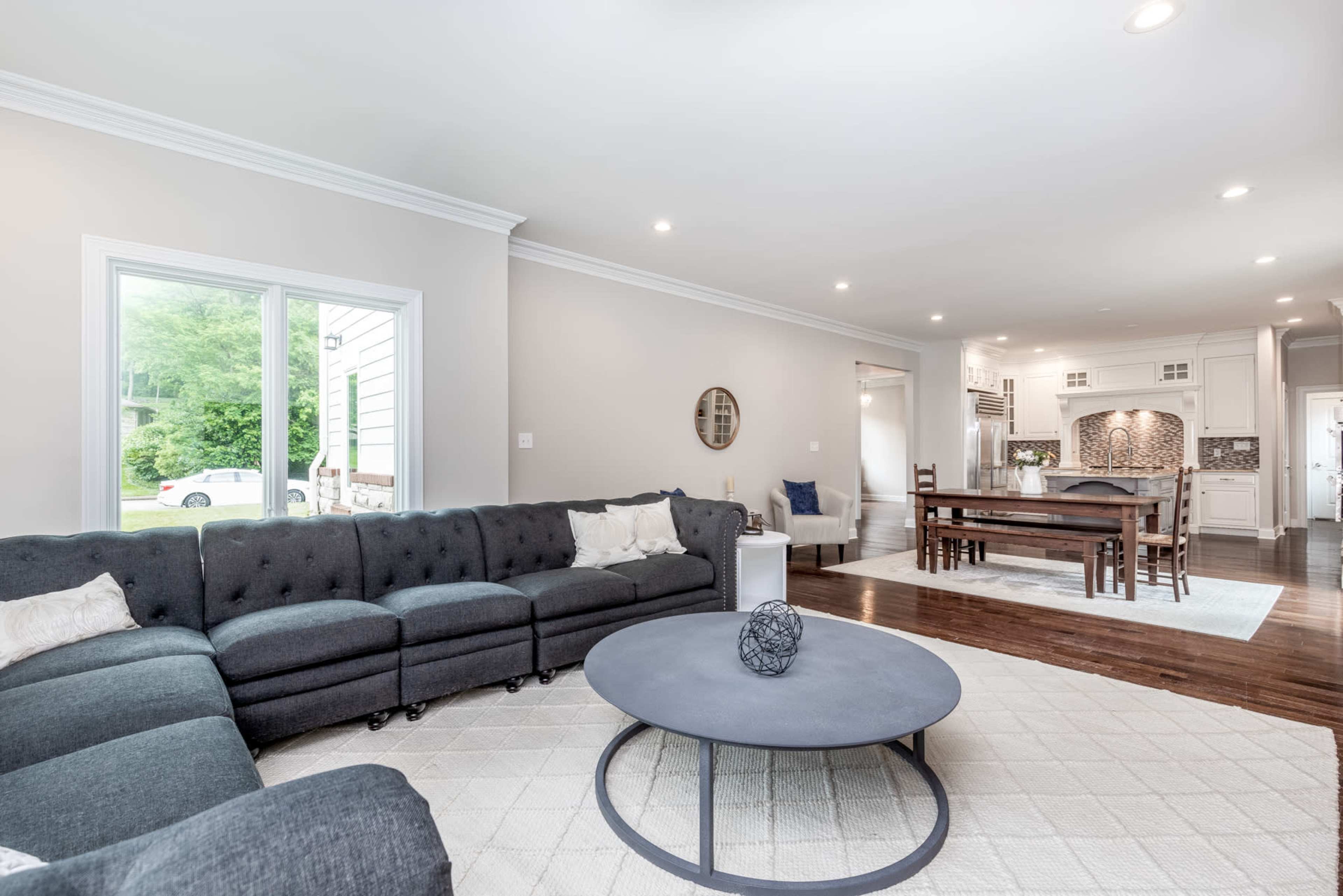 A well-lit living room featuring a gray sectional sofa, a round coffee table, and a view into a connected dining area with a wooden table.