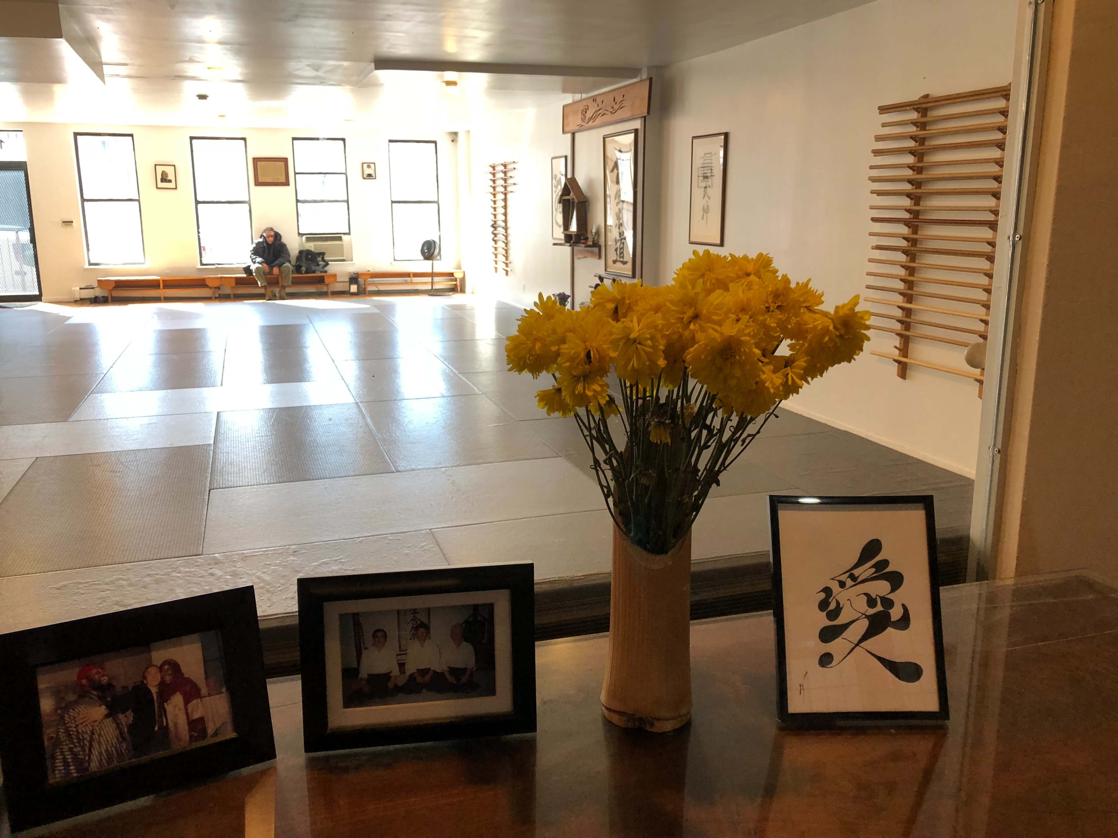 A spacious room with wooden benches and exercise equipment, featuring a table decorated with a vase of yellow flowers and framed photos.