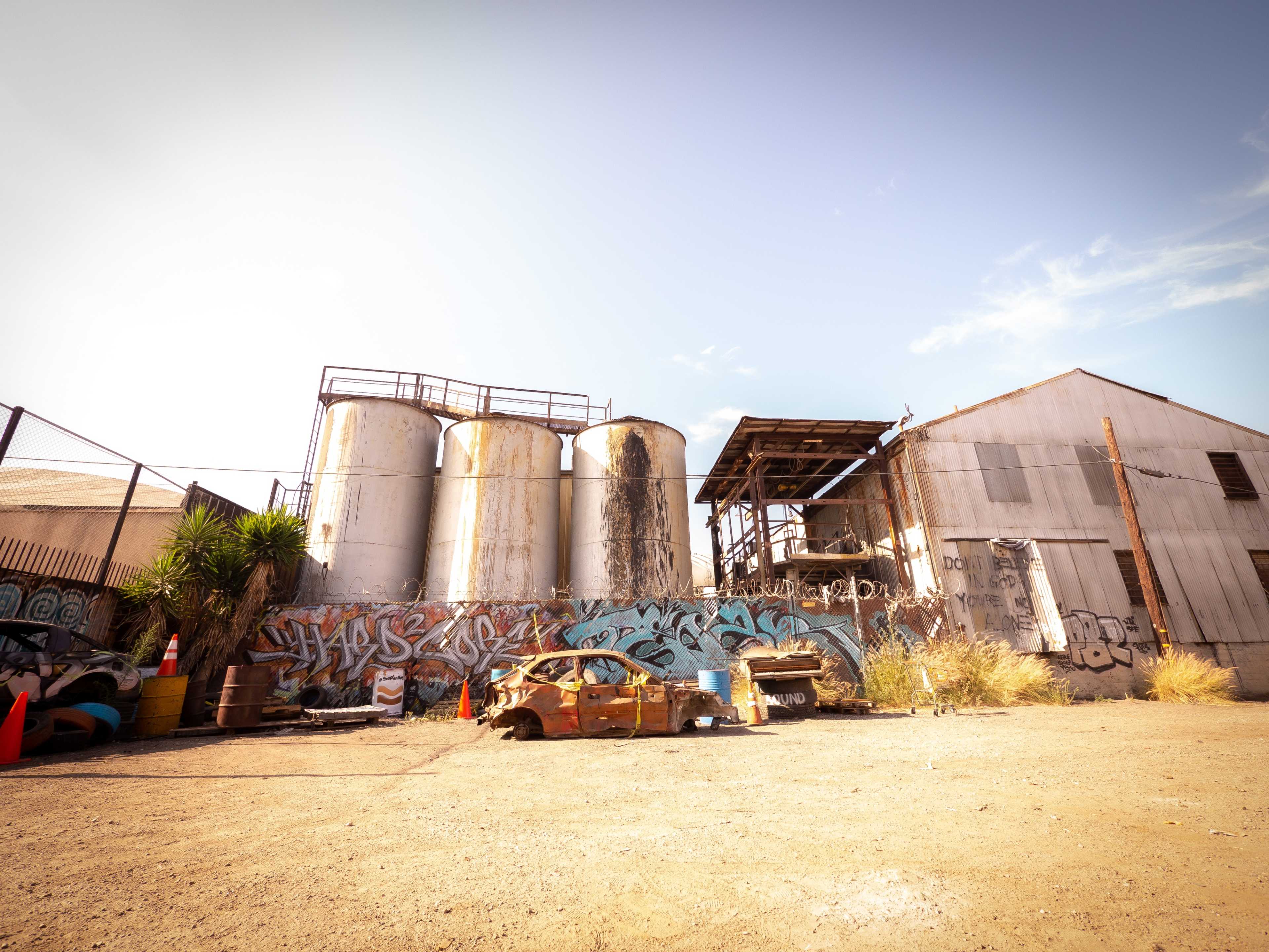 The image shows a deserted industrial area with three large silos, a dilapidated building, and a rusted car covered in graffiti.