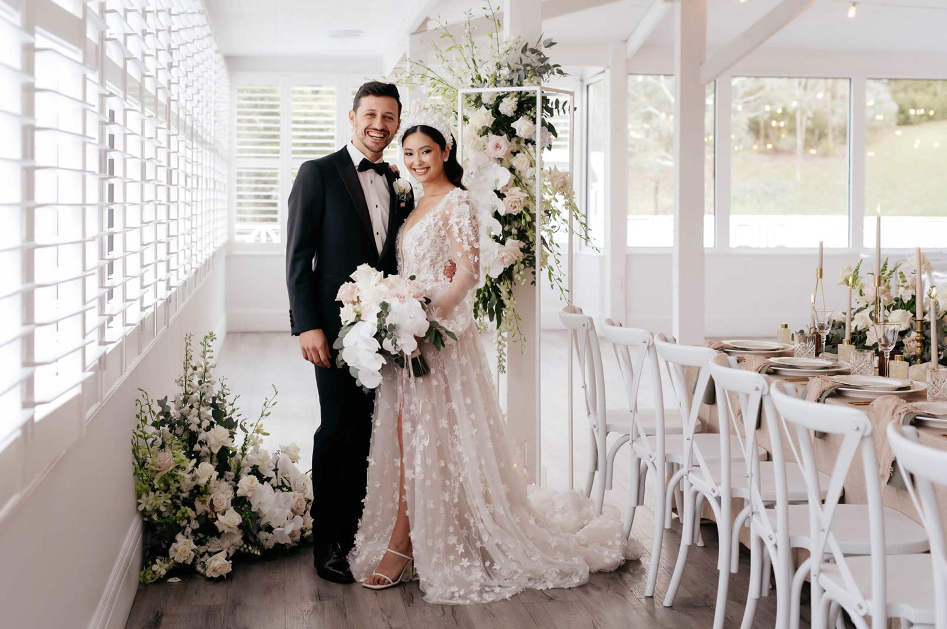 A couple stands together, smiling, in a bright venue decorated with flowers and elegant table settings for their wedding.
