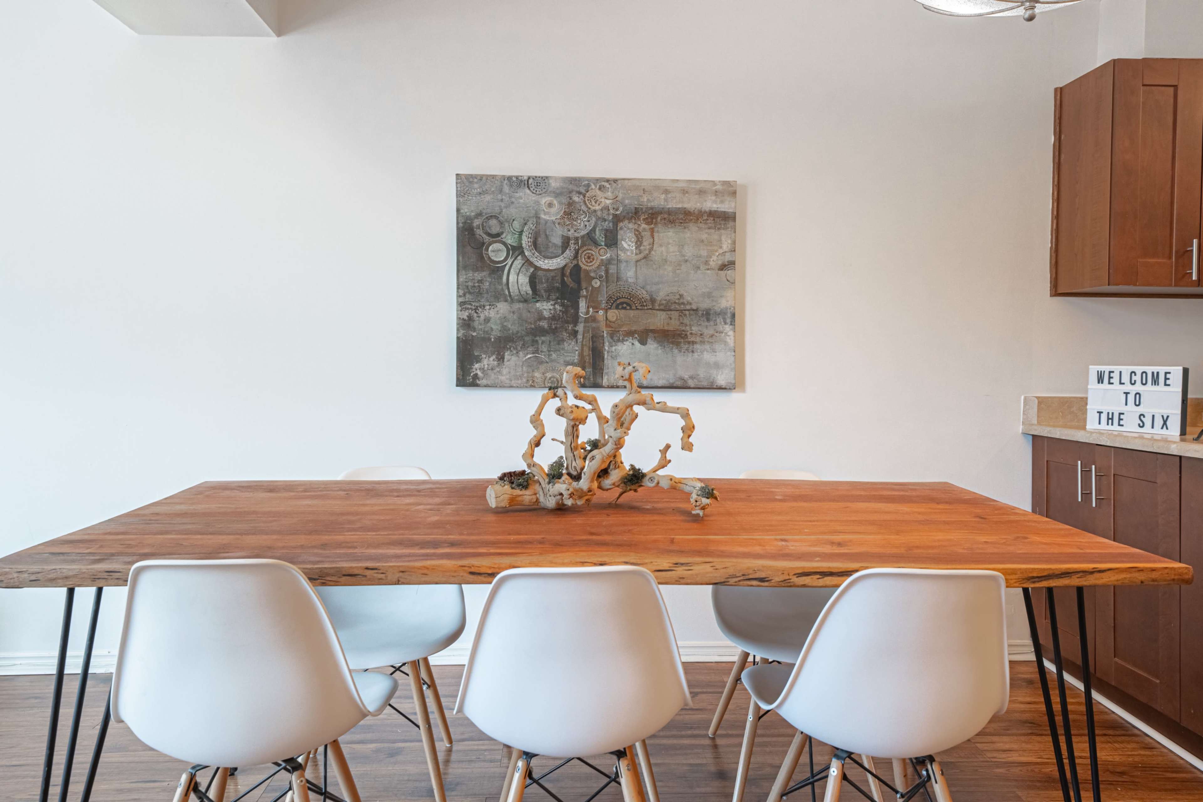 A wooden dining table with six white chairs is set against a wall featuring a modern artwork and a light-up sign that reads "WELCOME TO THE SIX."