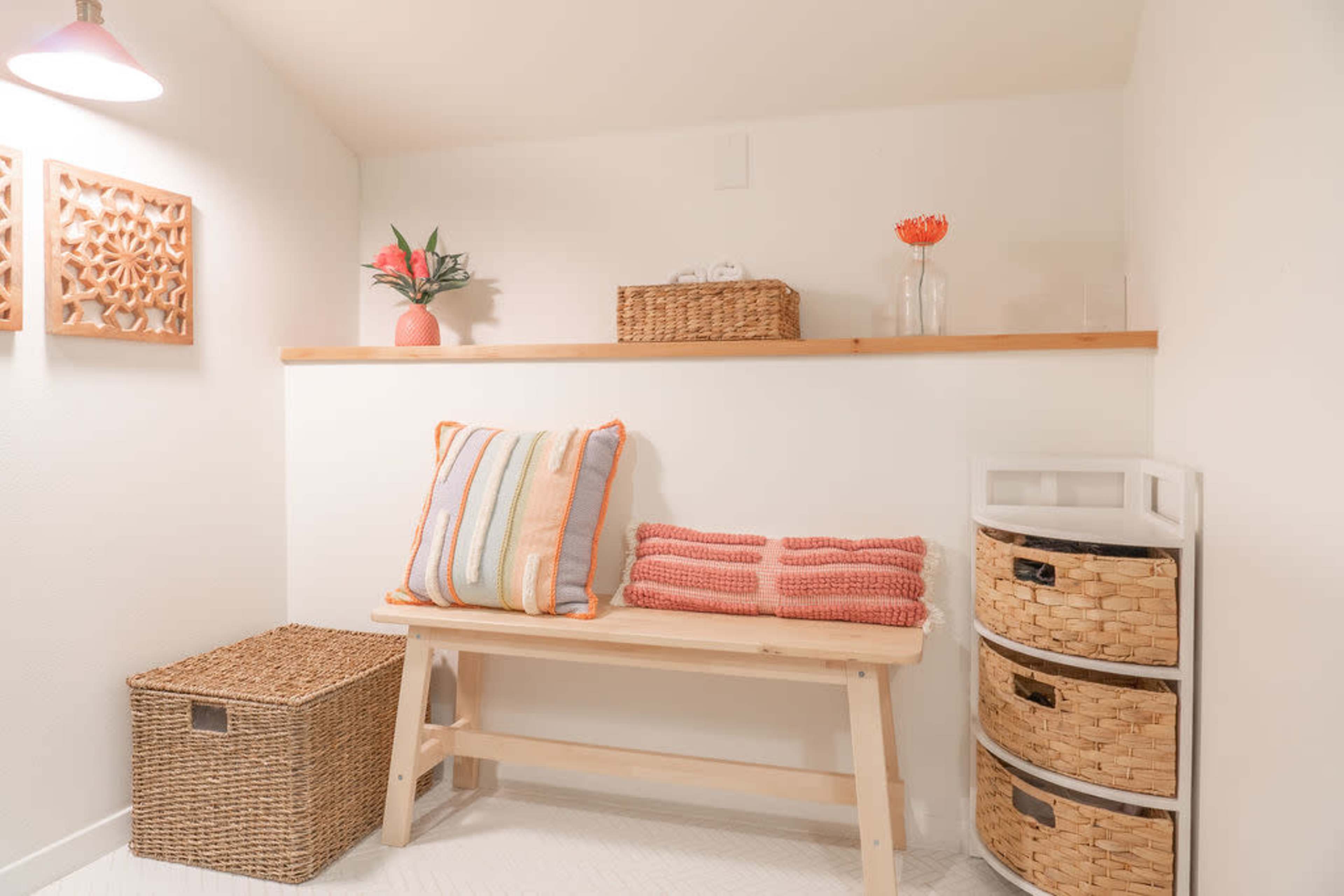 A small, organized space featuring a wooden bench with decorative cushions, a shelf with woven baskets, and a storage unit with drawers.