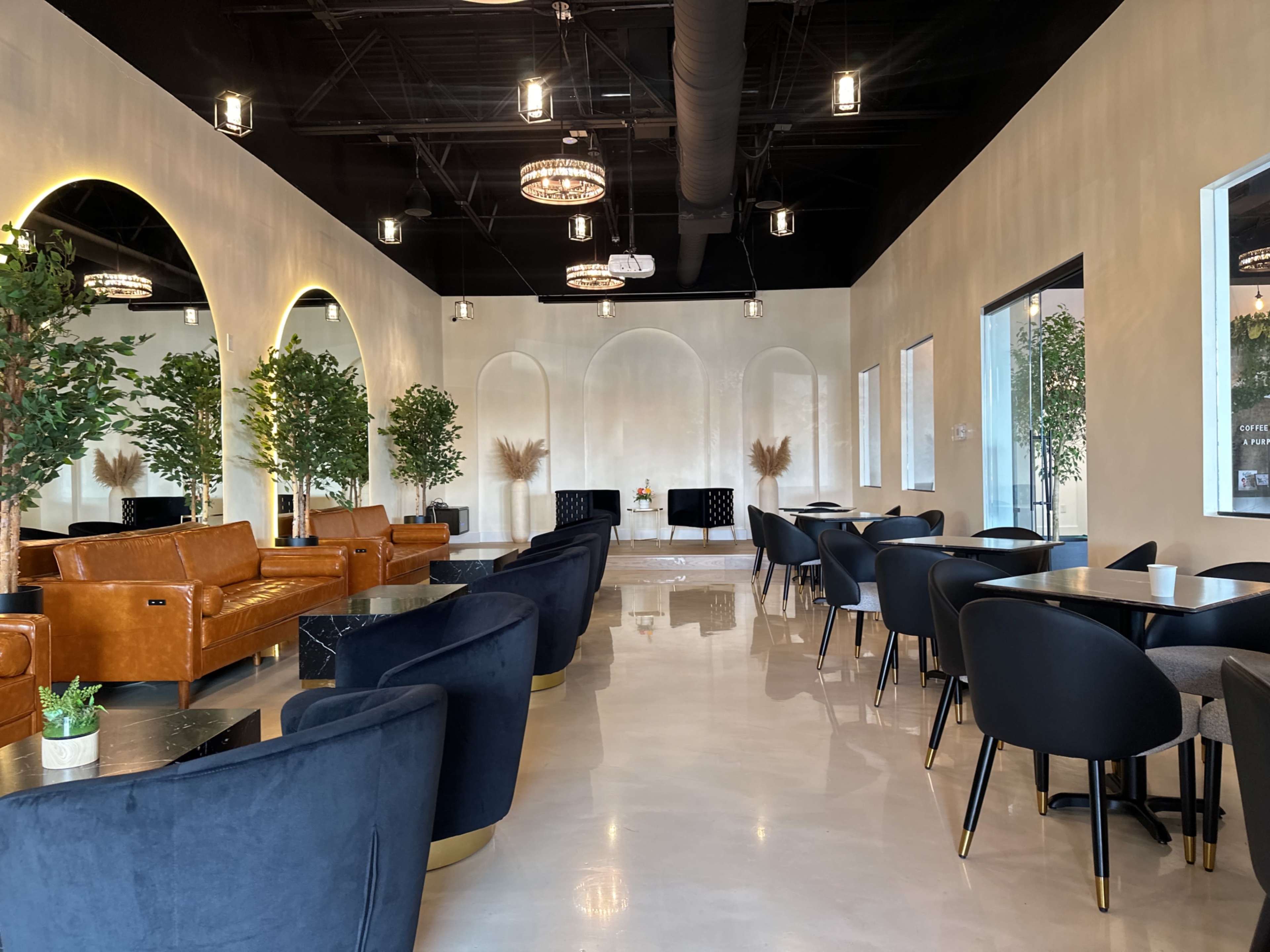 The image shows a modern café interior featuring brown leather sofas, round tables with black chairs, and decorative plants.