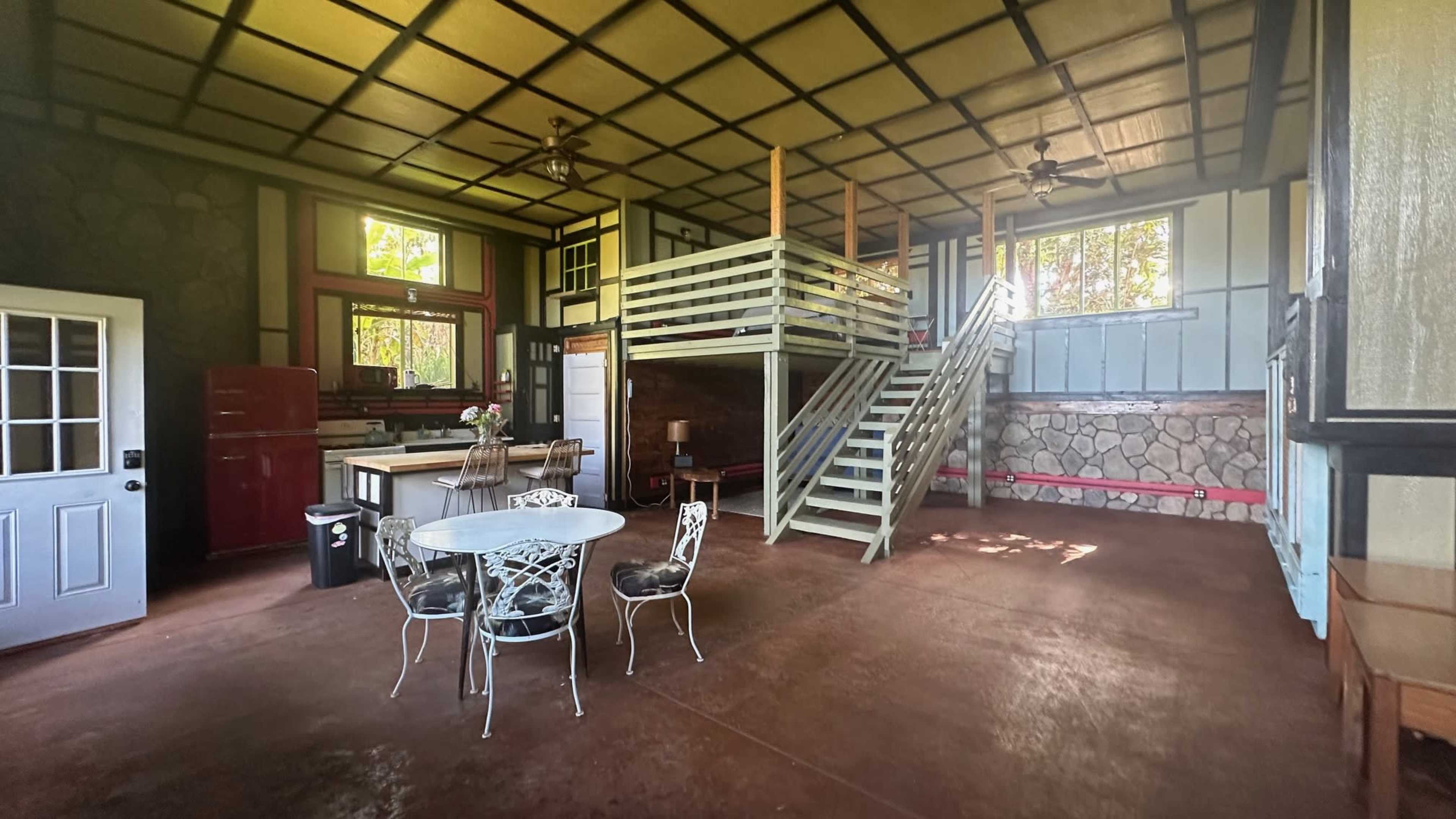 A spacious interior of a house featuring a dining area with a round table and chairs, a staircase leading to a loft, and a kitchen area in the background.