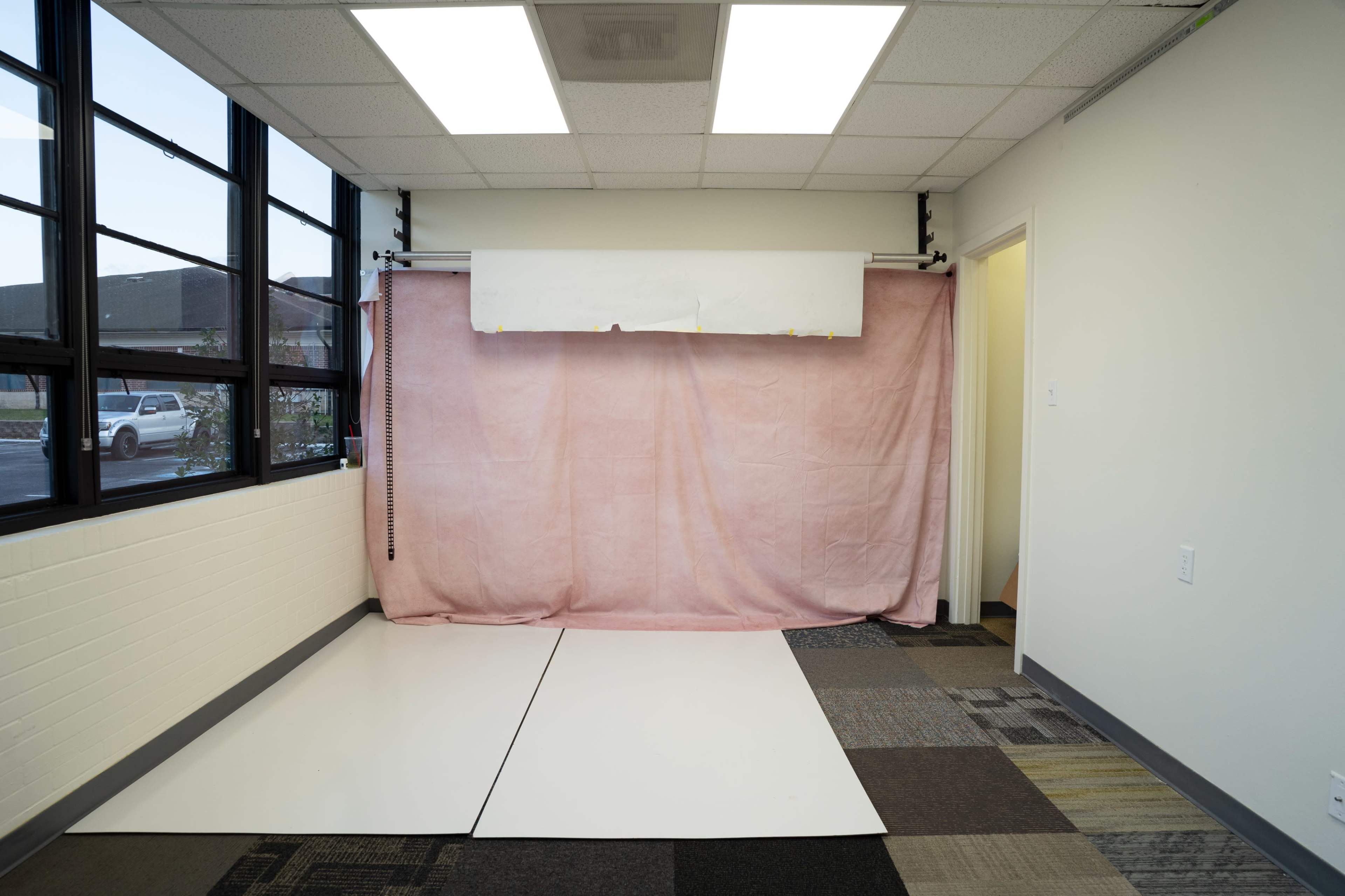 The image shows a simple room featuring a pink backdrop draped on a wall, with white flooring and large windows allowing natural light.