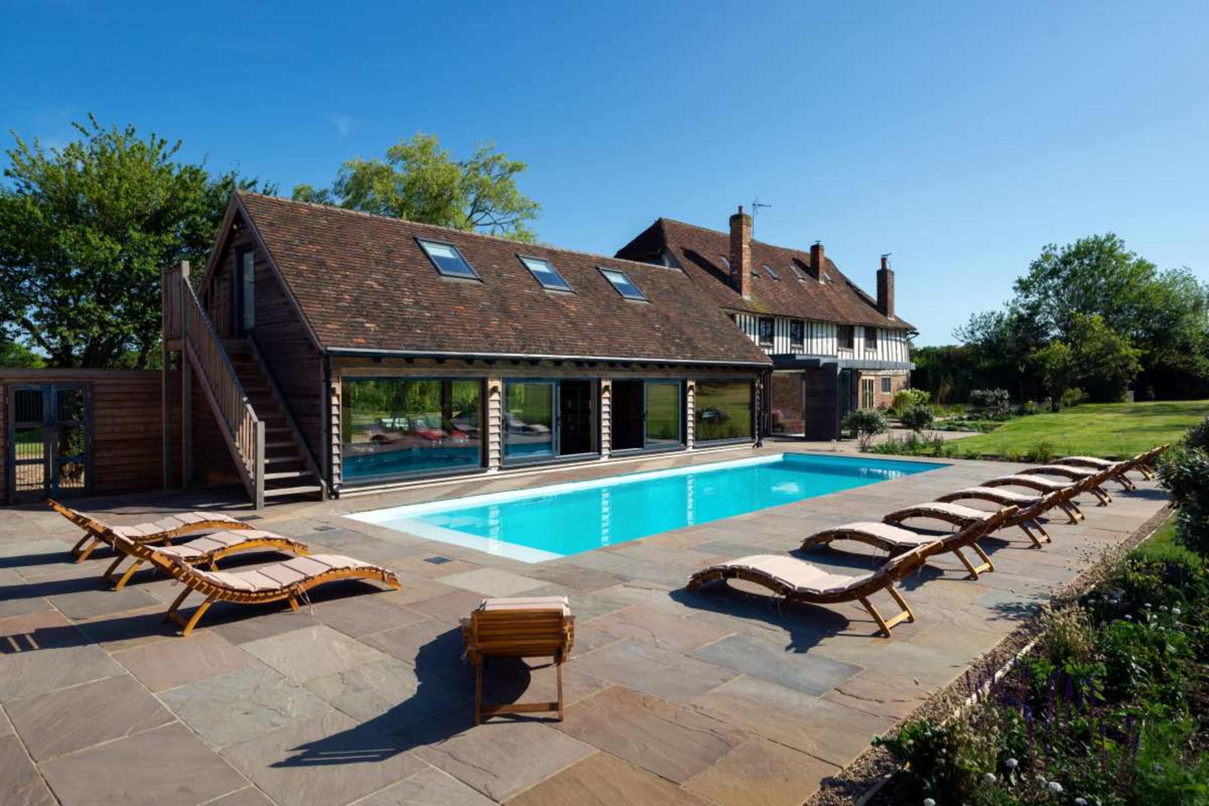 A modern house with large windows overlooks a pool surrounded by lounge chairs on a stone patio.