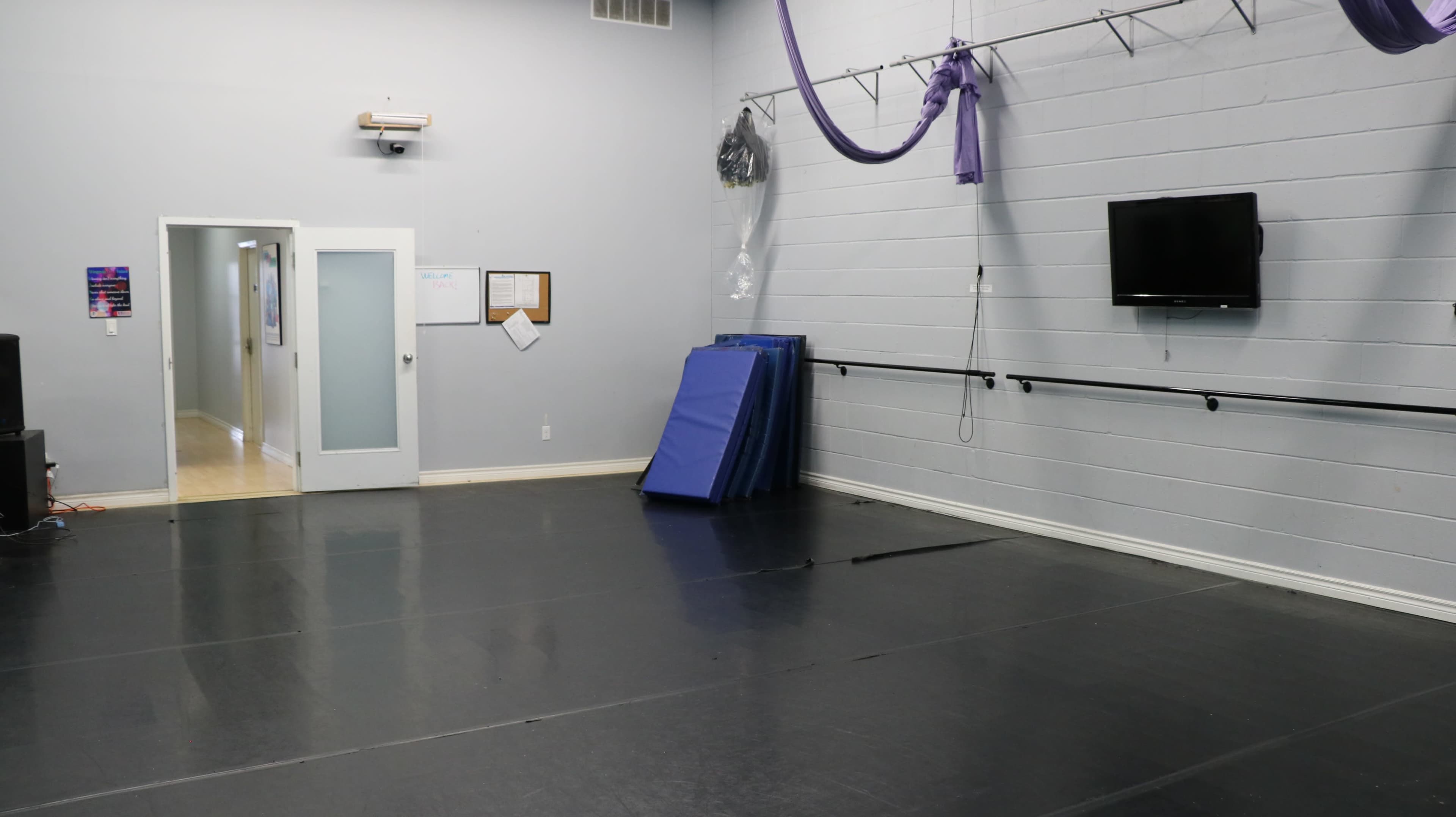 The image shows an empty dance studio with a mirrored wall, black flooring, a television mounted on the wall, and stacked blue mats.