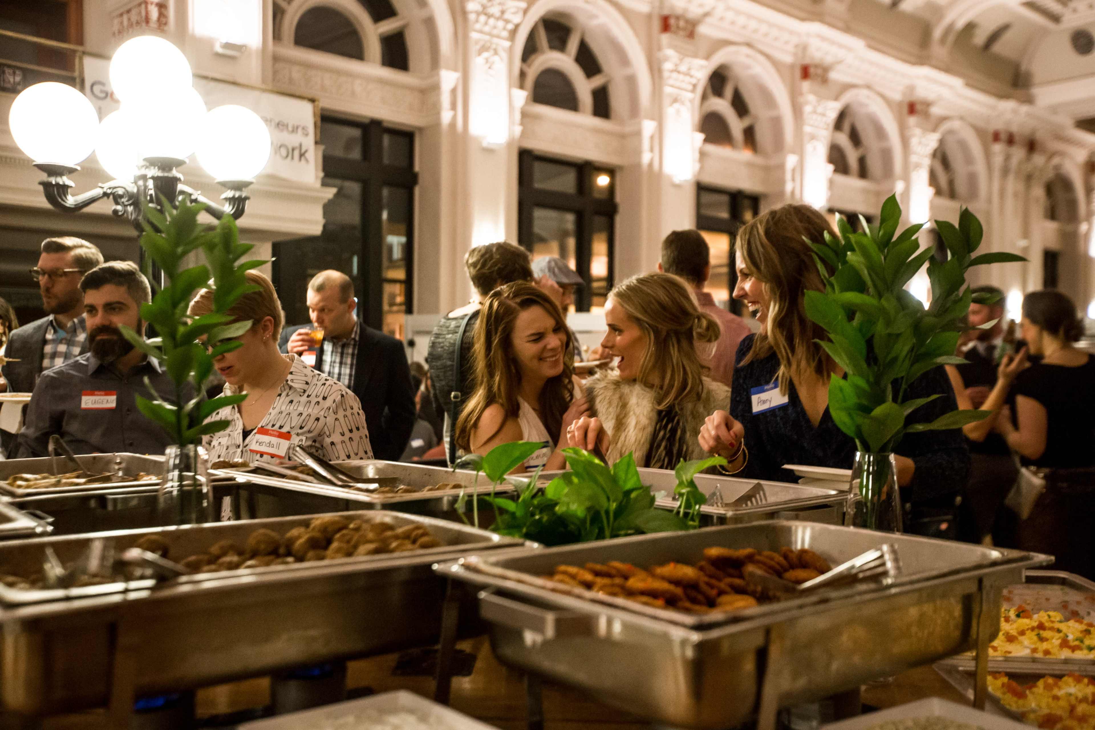 A group of people socializes at a buffet table filled with various food items in a well-lit indoor space.