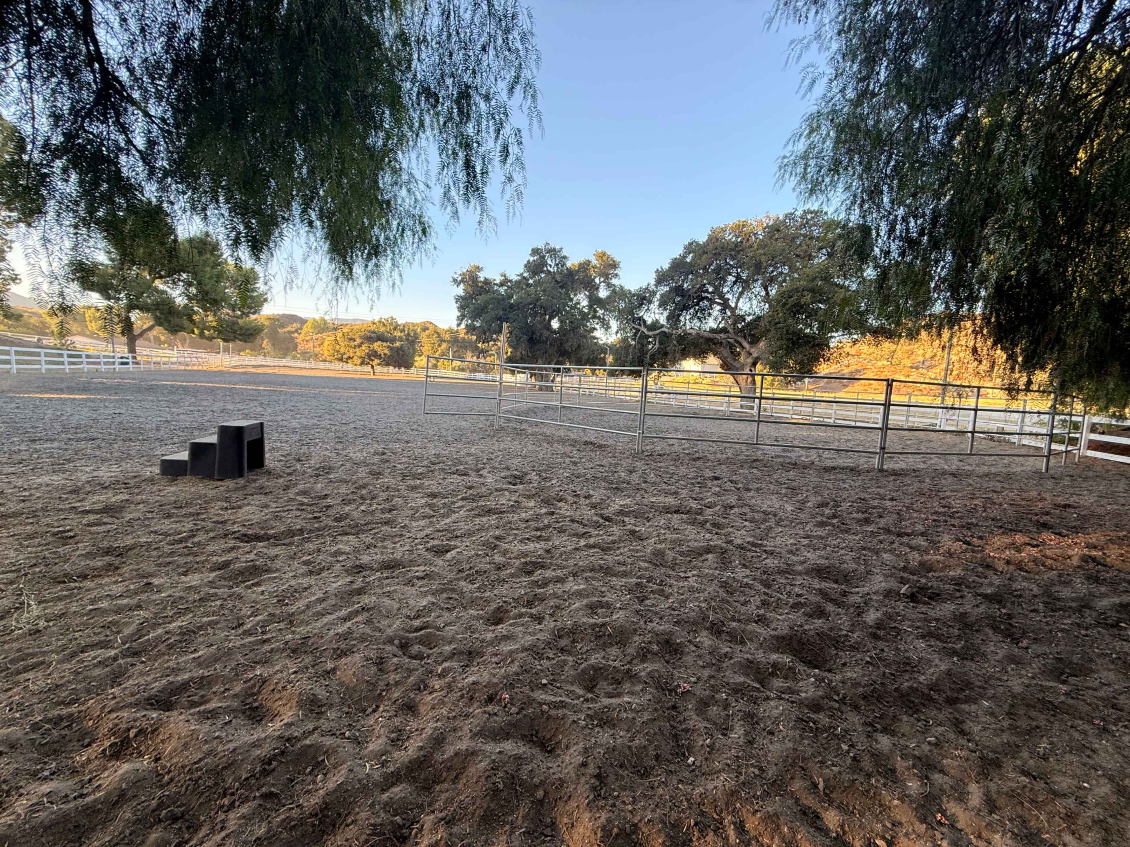 The image shows a fenced, sandy area with sparse vegetation and a few black boxes in one corner, surrounded by trees and an open field in the background.