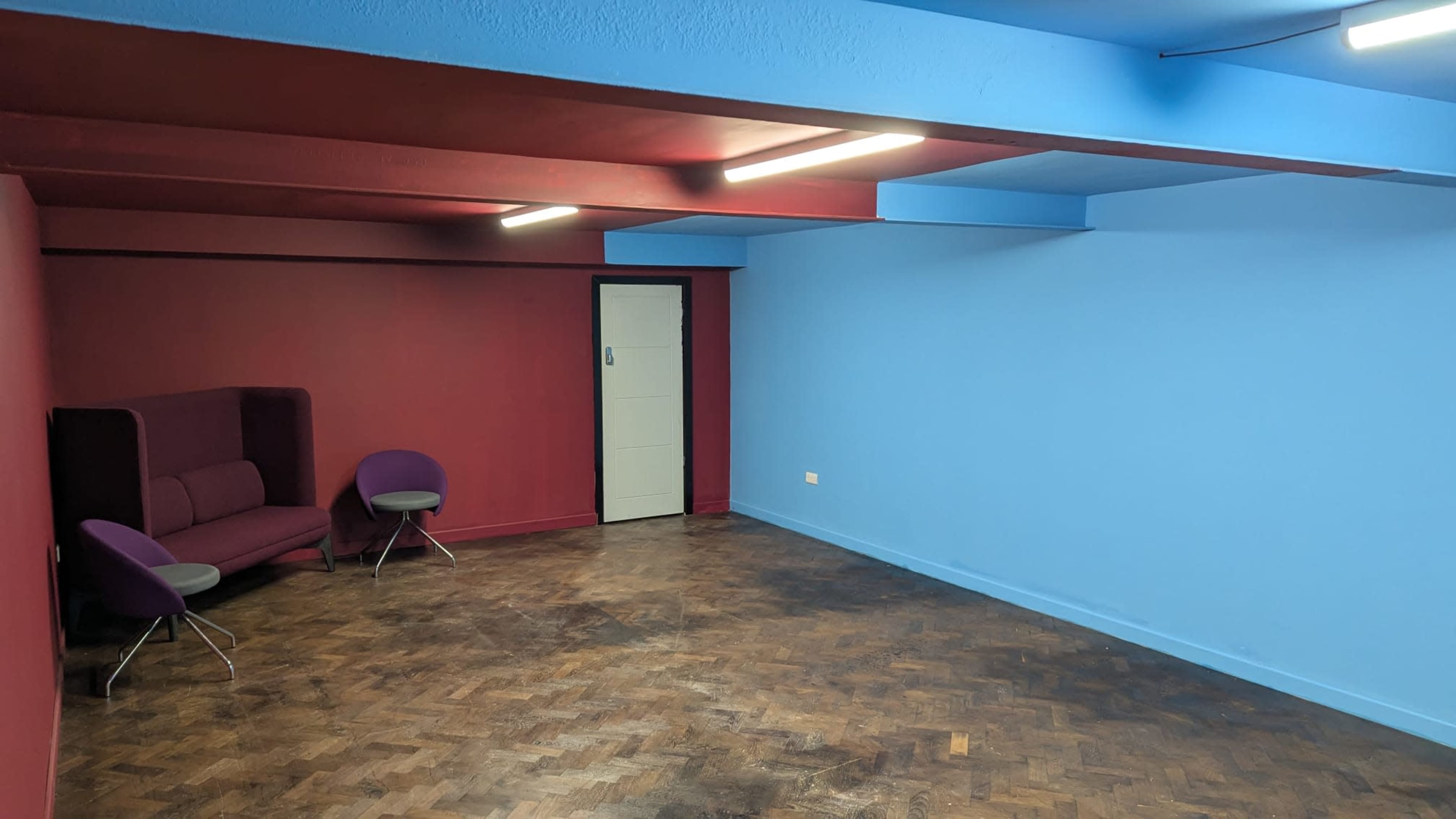 The image shows an empty room with a blue and red color scheme, featuring a dark wooden floor and a small seating area with two purple chairs.
