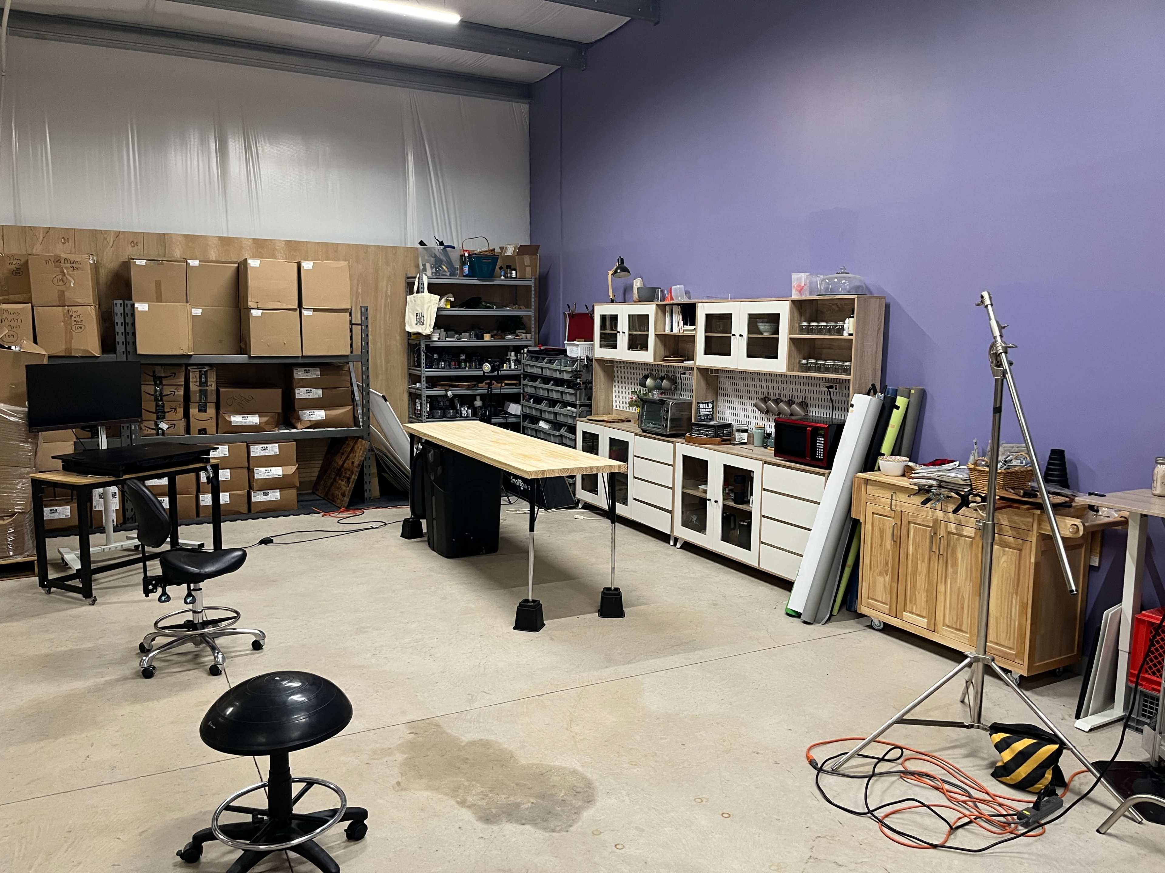 A work area with a wooden table in the center, surrounded by storage shelves, a computer station, and various tools and equipment.