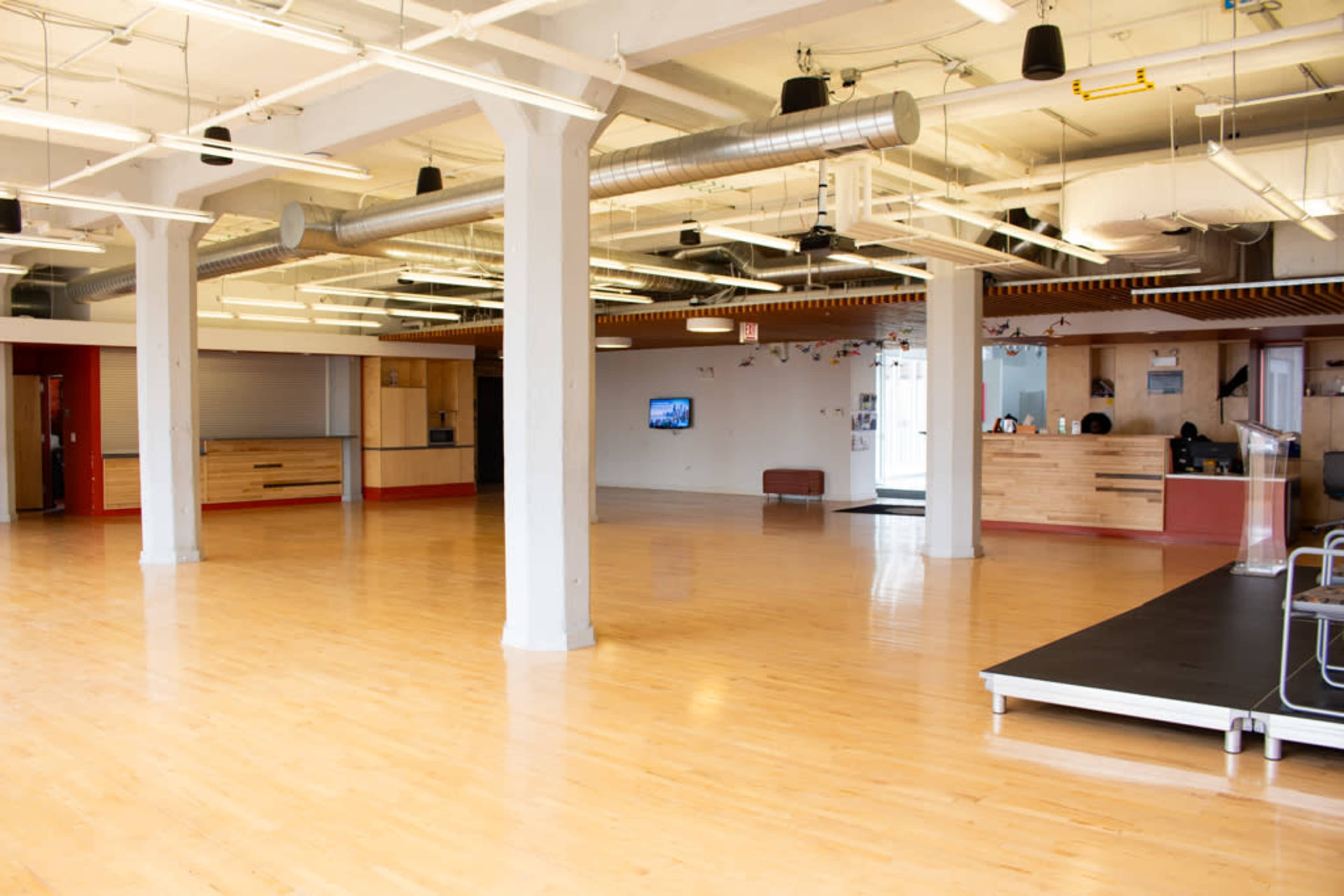 The image shows a spacious, well-lit interior with wooden flooring, exposed beams, and minimal furnishings.