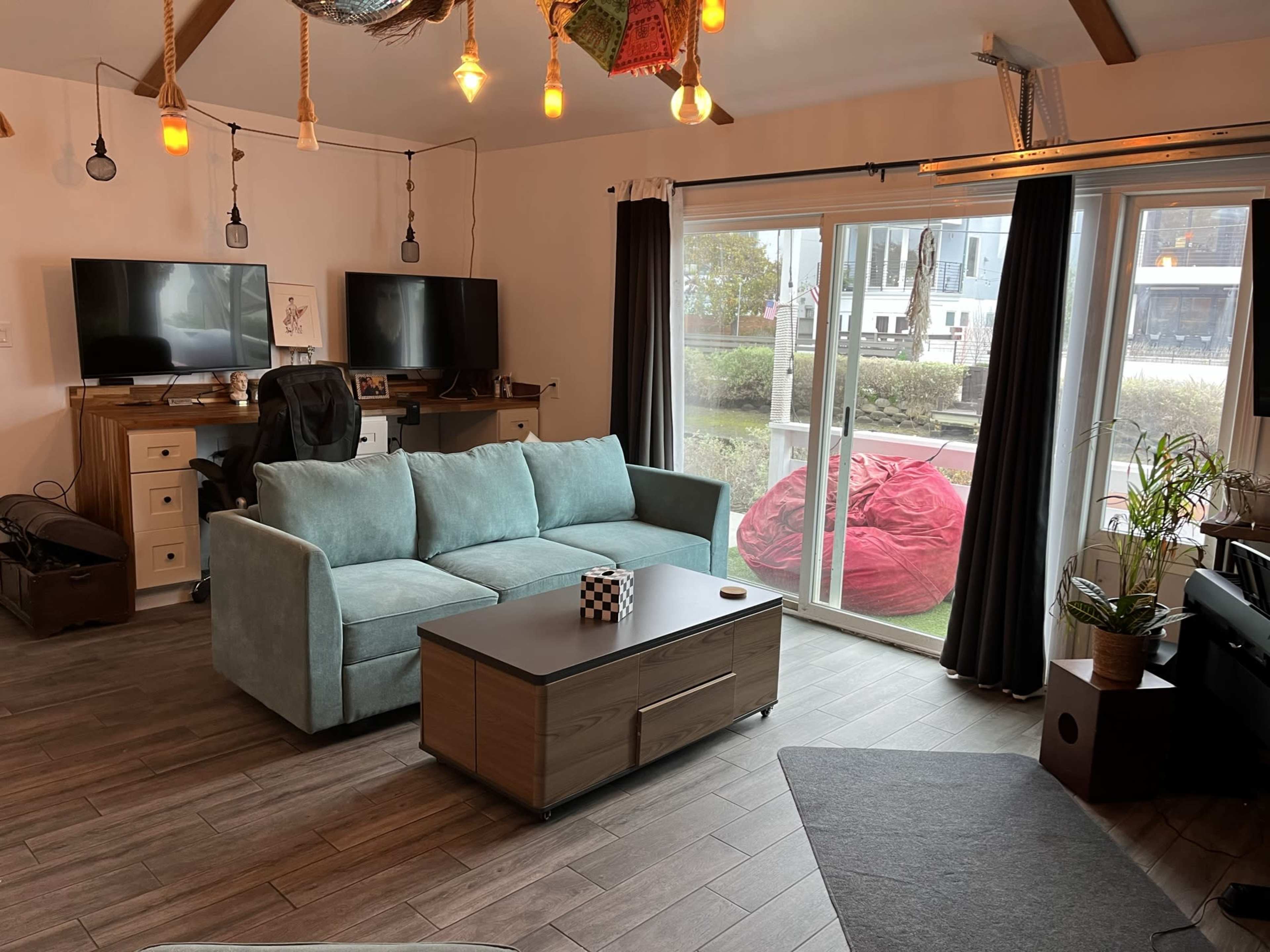A living room with a light blue couch, a wooden coffee table, two television sets on a wall unit, and large sliding glass doors leading to an outdoor area filled with greenery.