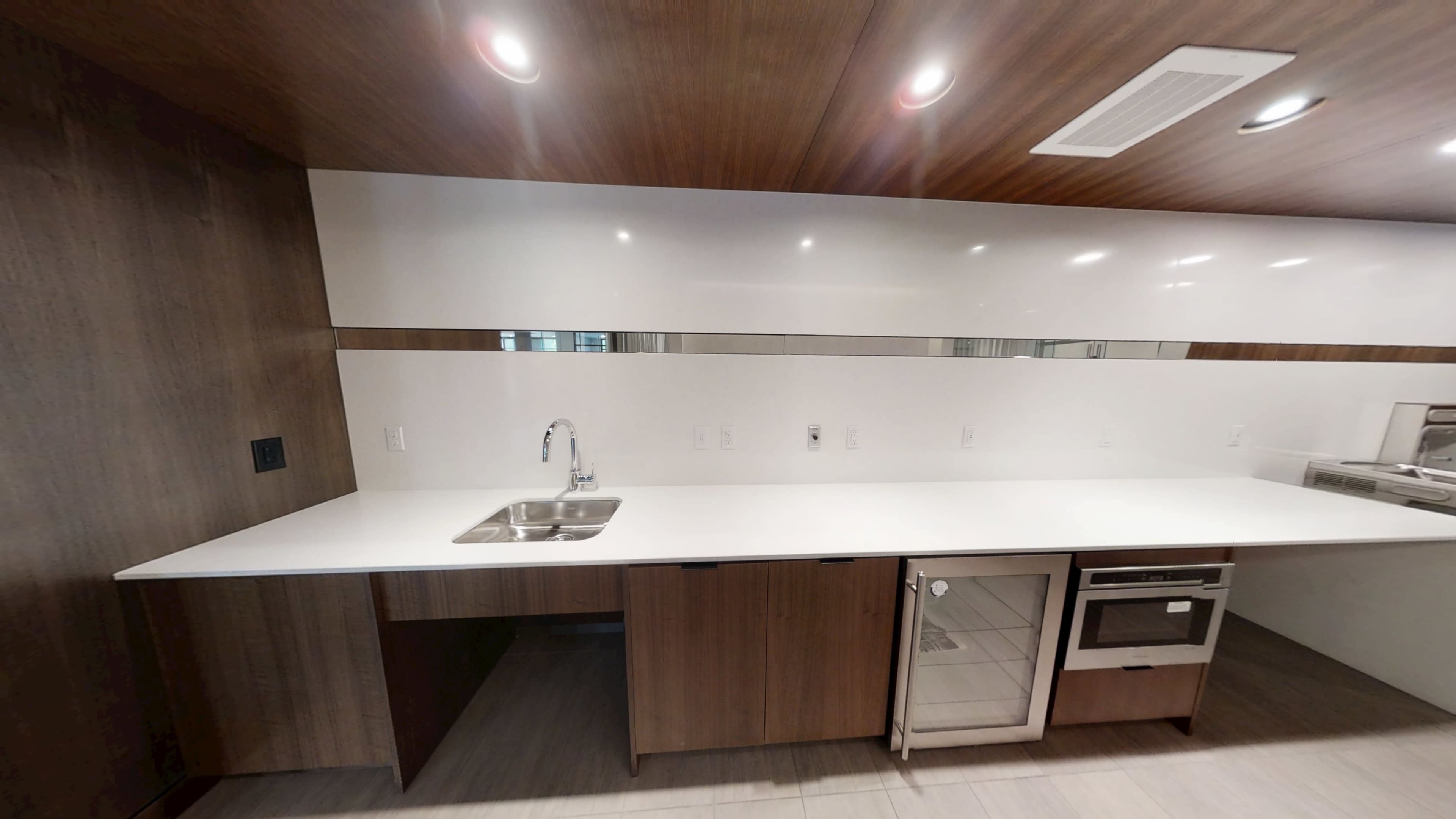 The image shows a modern kitchen with a white countertop, a sink, and built-in appliances set against wooden cabinetry and a glossy wall.