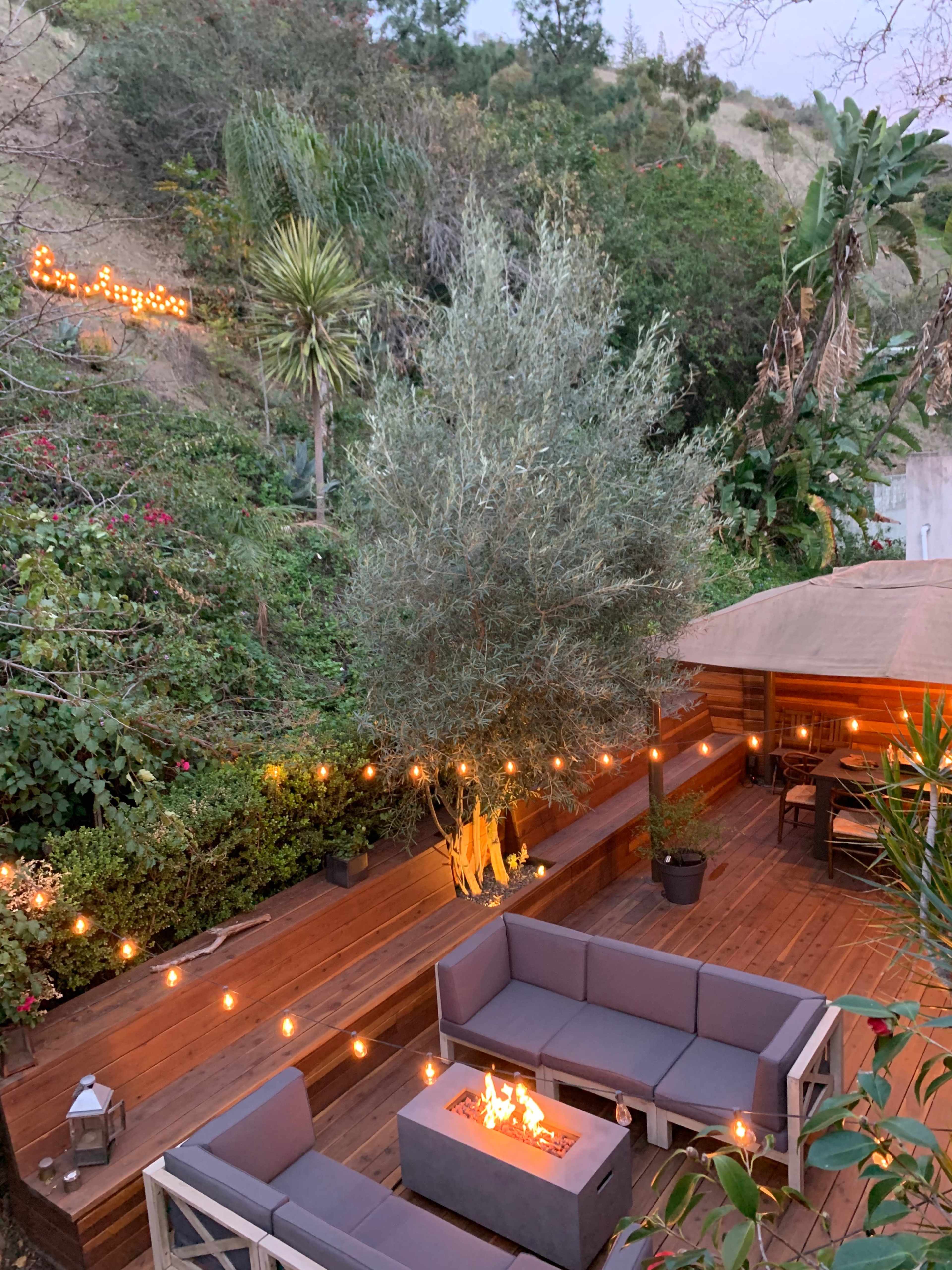 The image shows a wooden deck with a fire pit surrounded by outdoor furniture and string lights, set amidst lush greenery.