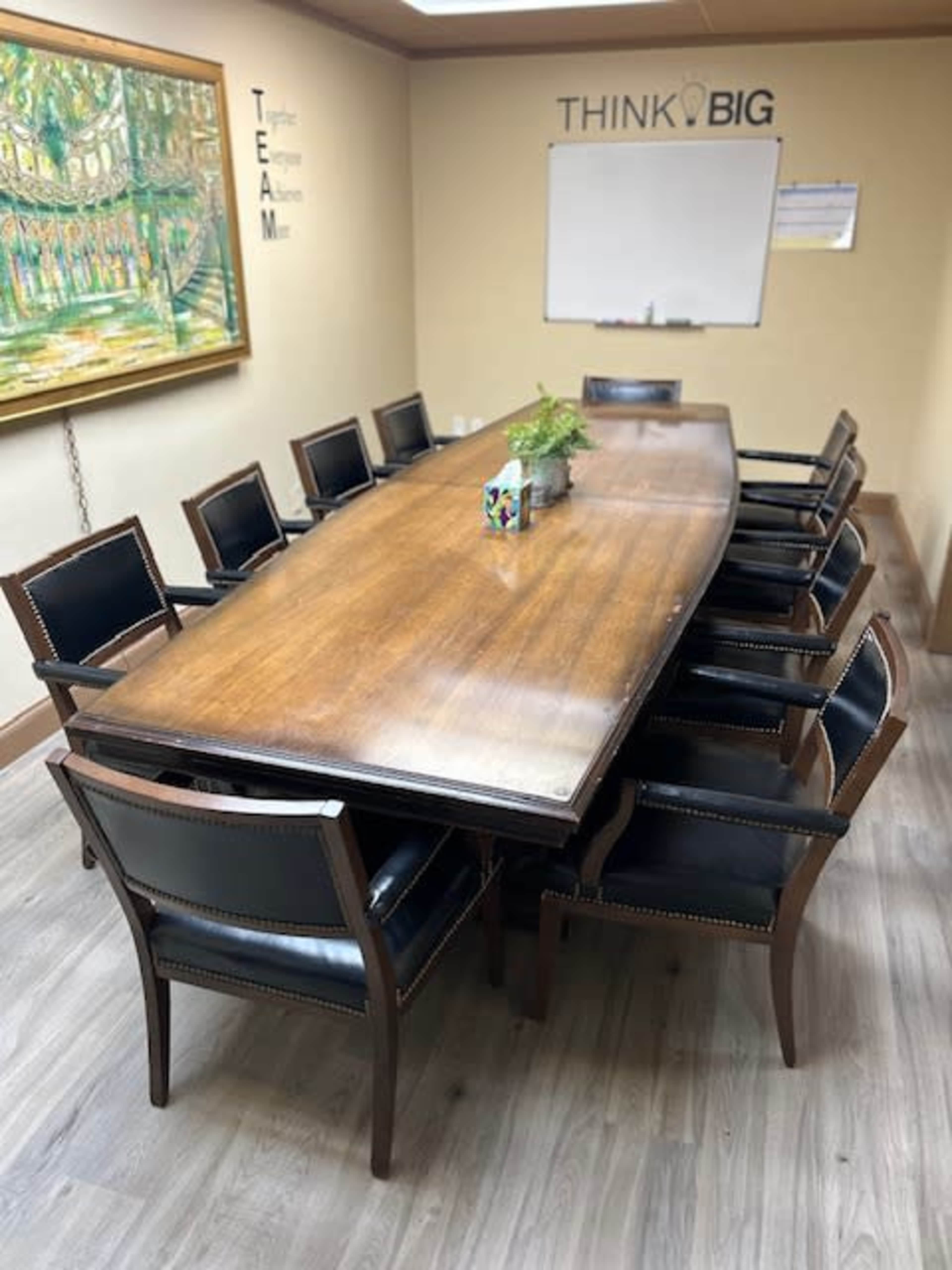 A long wooden conference table surrounded by black leather chairs is set in a meeting room with a whiteboard and a decorative painting on the wall.