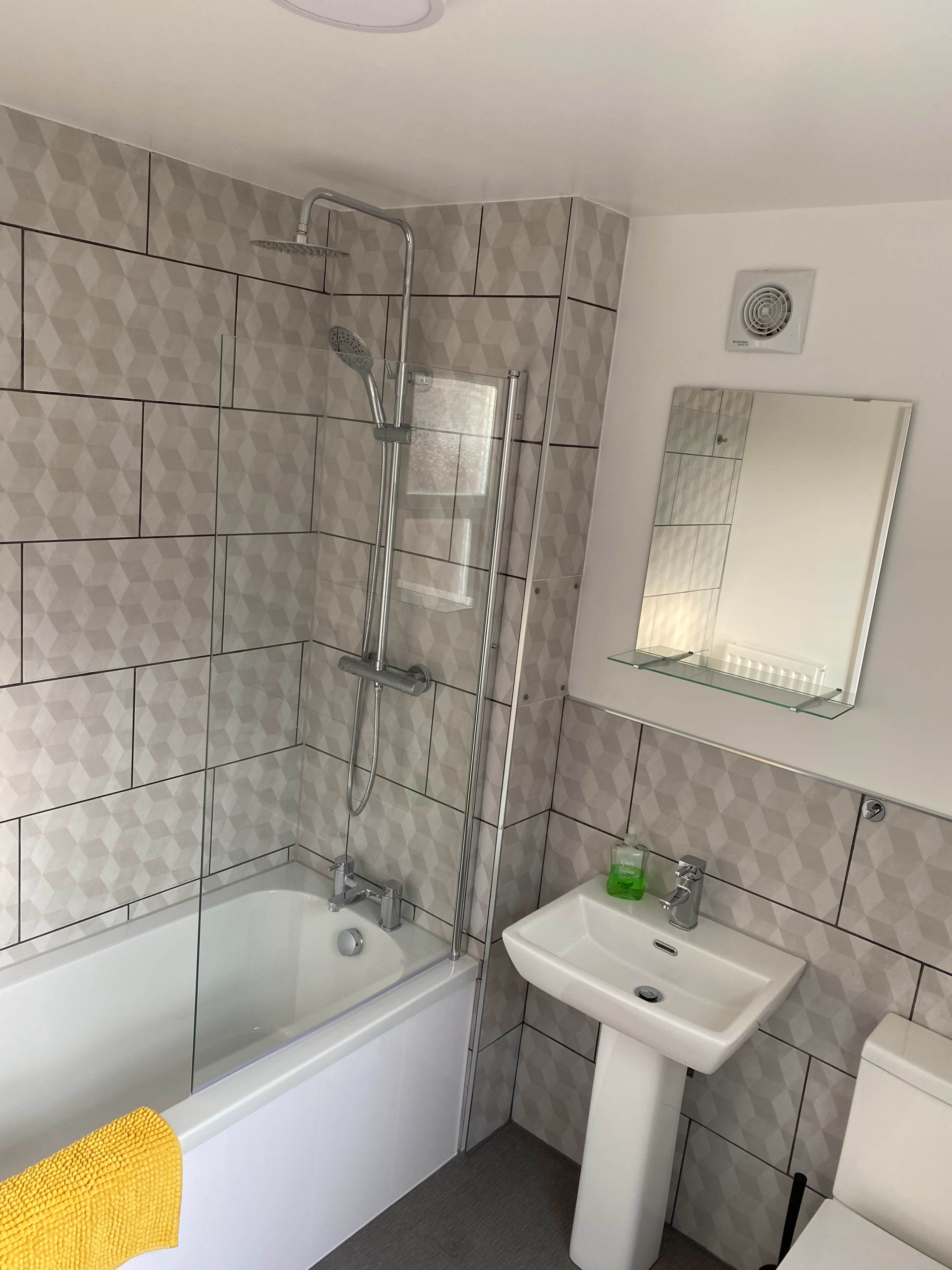 The image shows a bathroom featuring a bathtub with a shower, a sink with a mirror above it, and tiled walls with a geometric pattern.