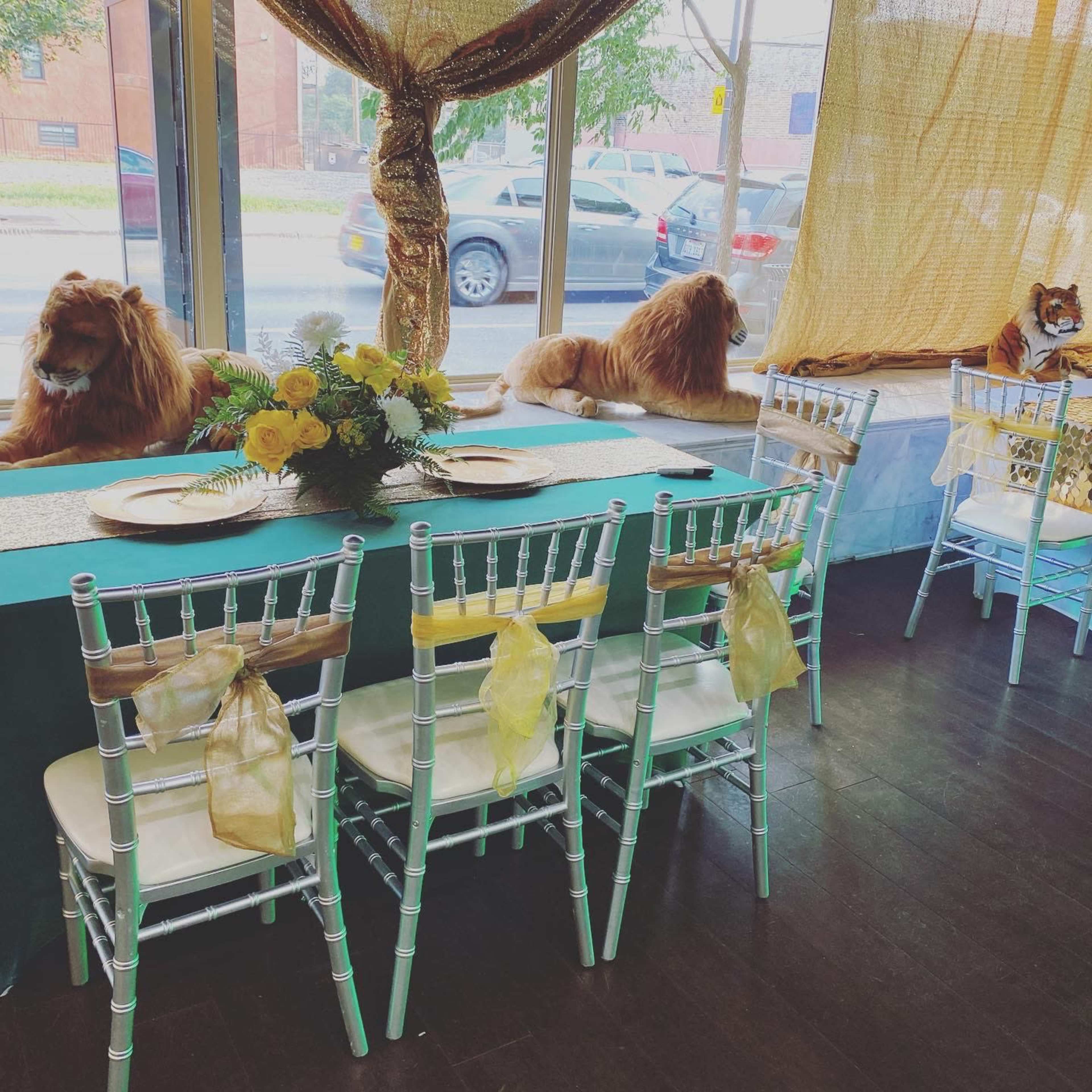 The image shows a dining setup in a decorated interior with several chairs and a table adorned with flowers, while two lifelike lion statues sit in the background.
