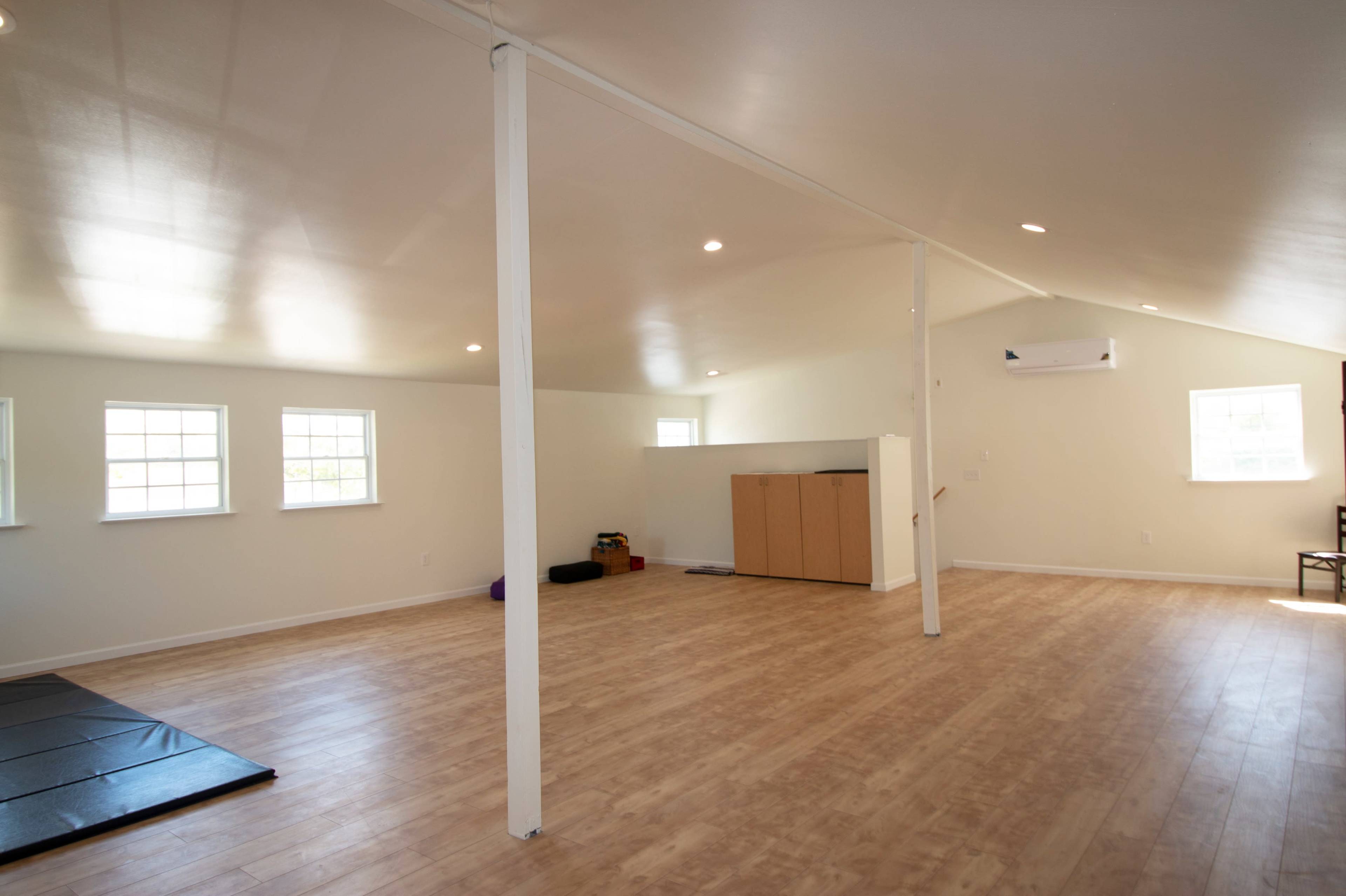 The image shows a spacious, empty room with wooden floors, large windows, and a storage unit against the wall.
