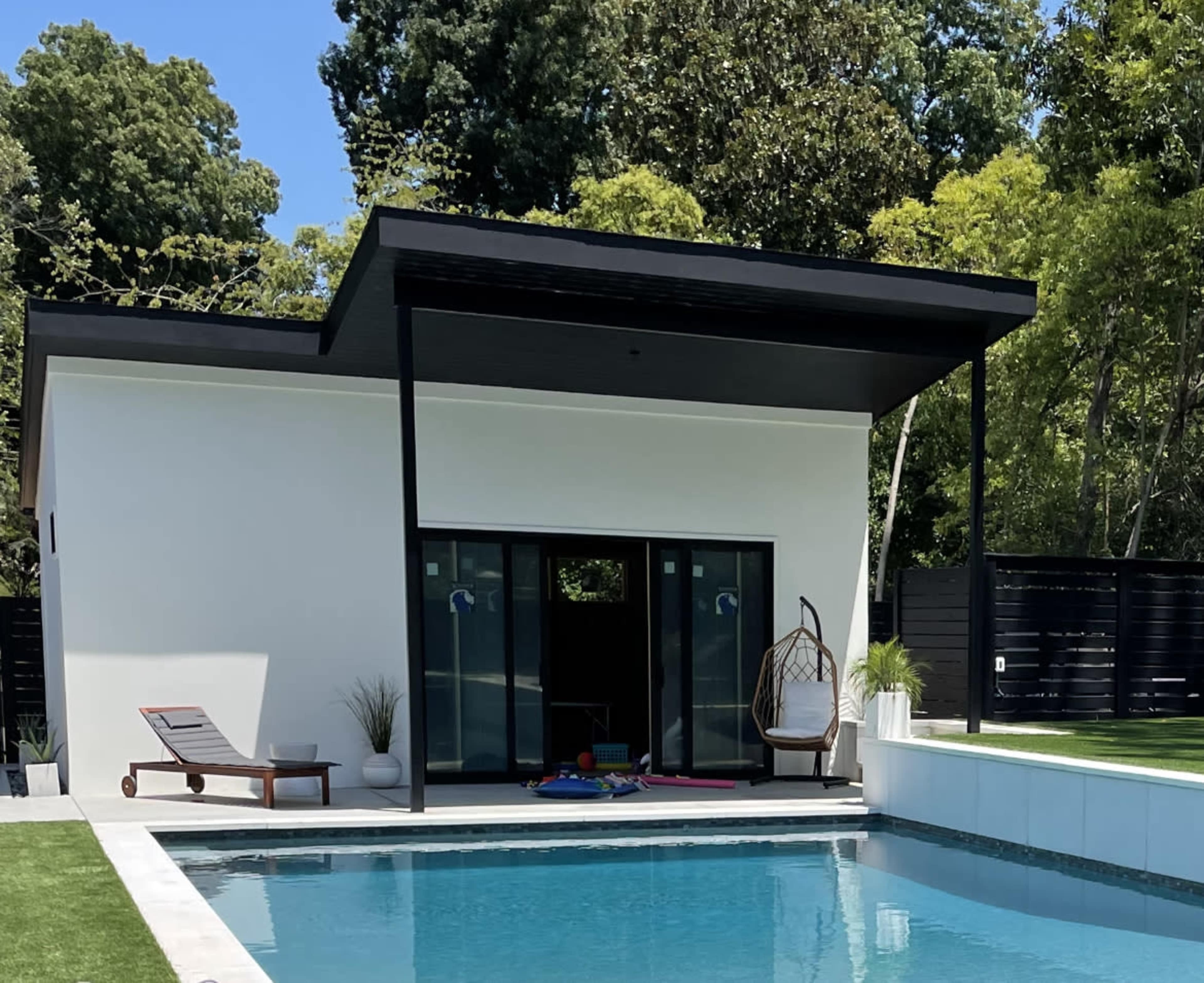 A modern poolside structure with a flat roof overlooks a clear blue pool, featuring large glass doors and a small patio area with lounge chairs and a hanging chair.