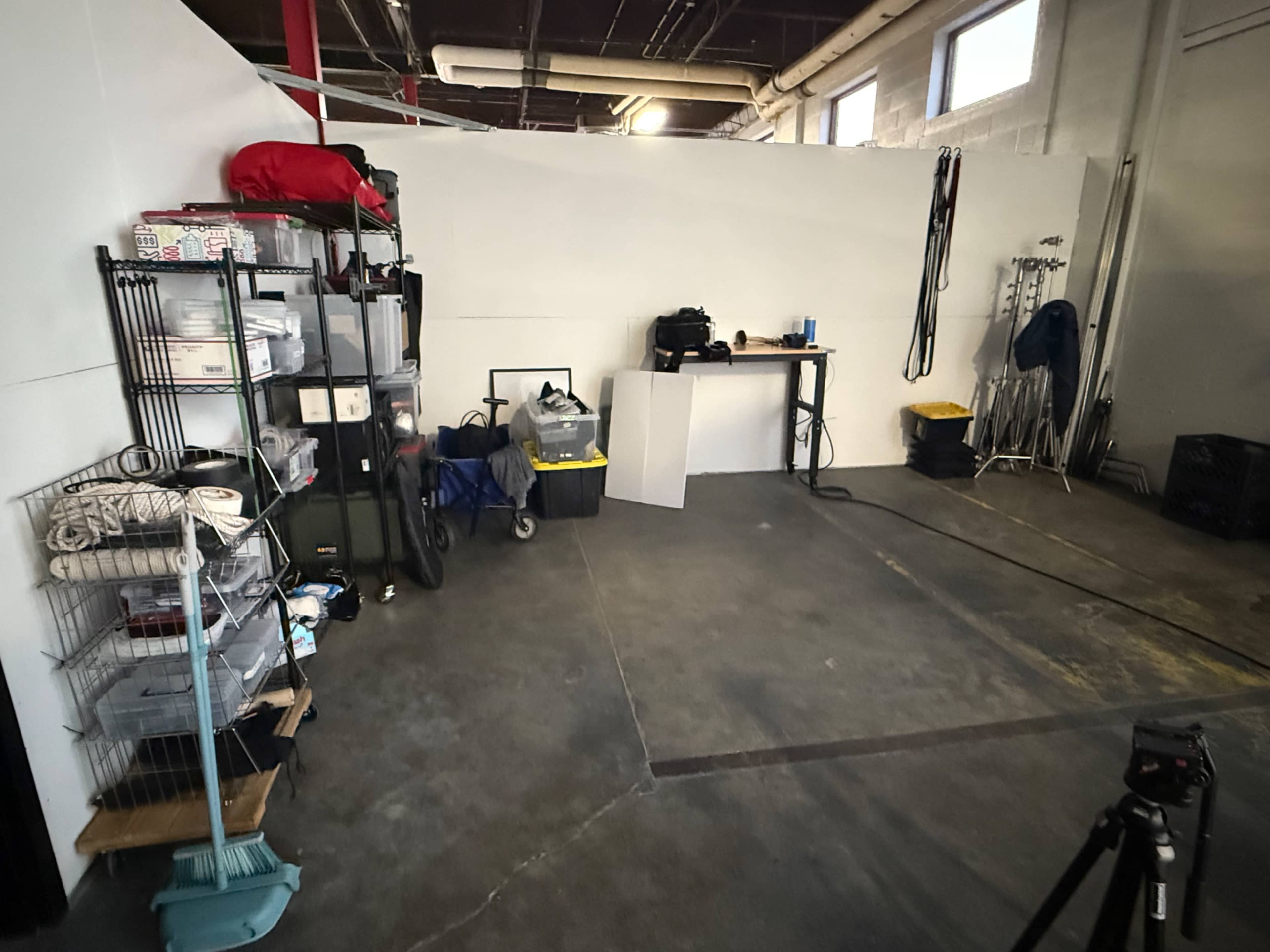 The image shows a room with various storage items, including a shelving unit, a table, and equipment scattered in a mostly empty space with gray flooring.