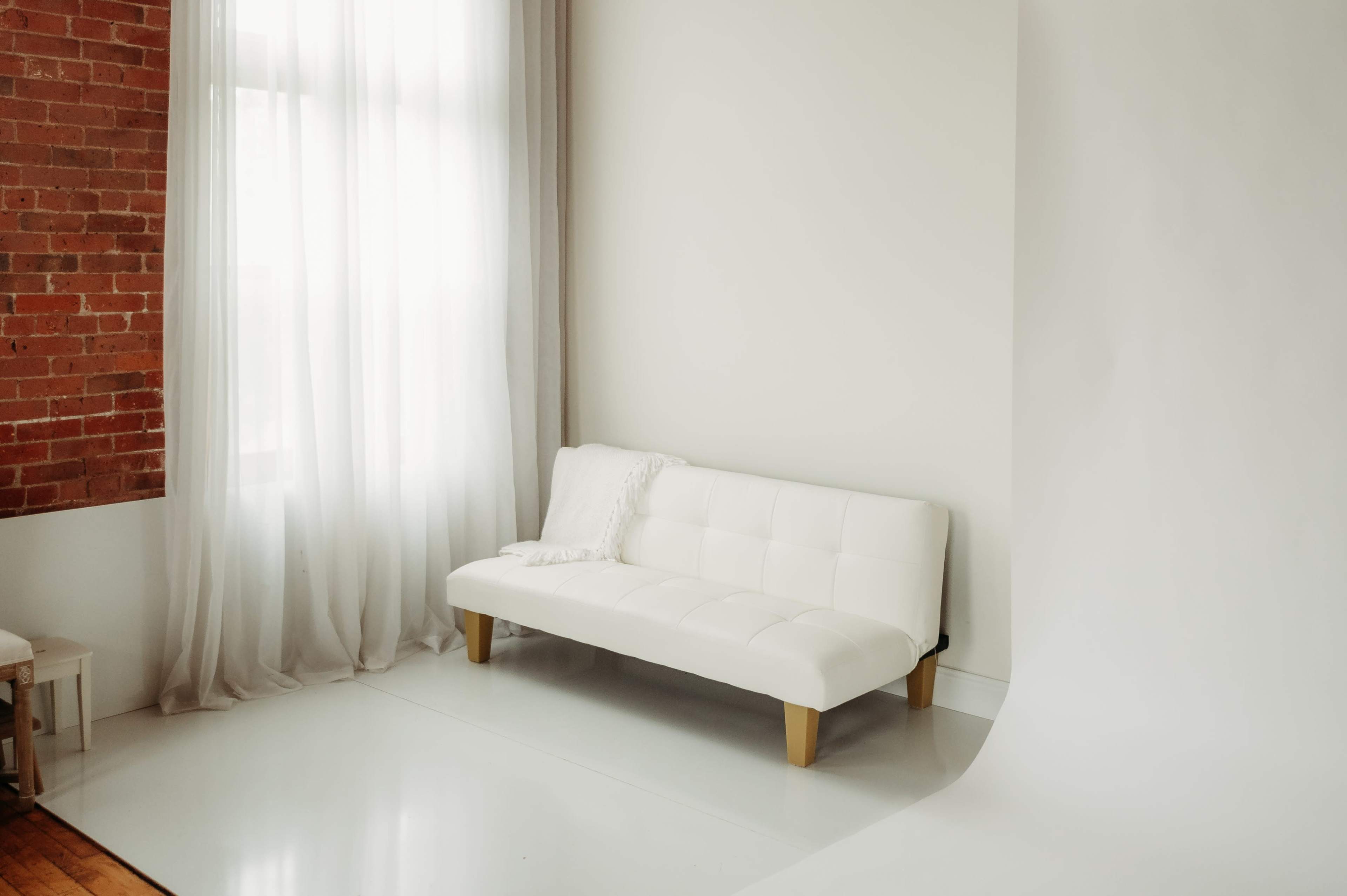A minimalistic interior featuring a white futon sofa against a plain wall with sheer curtains and exposed brick.