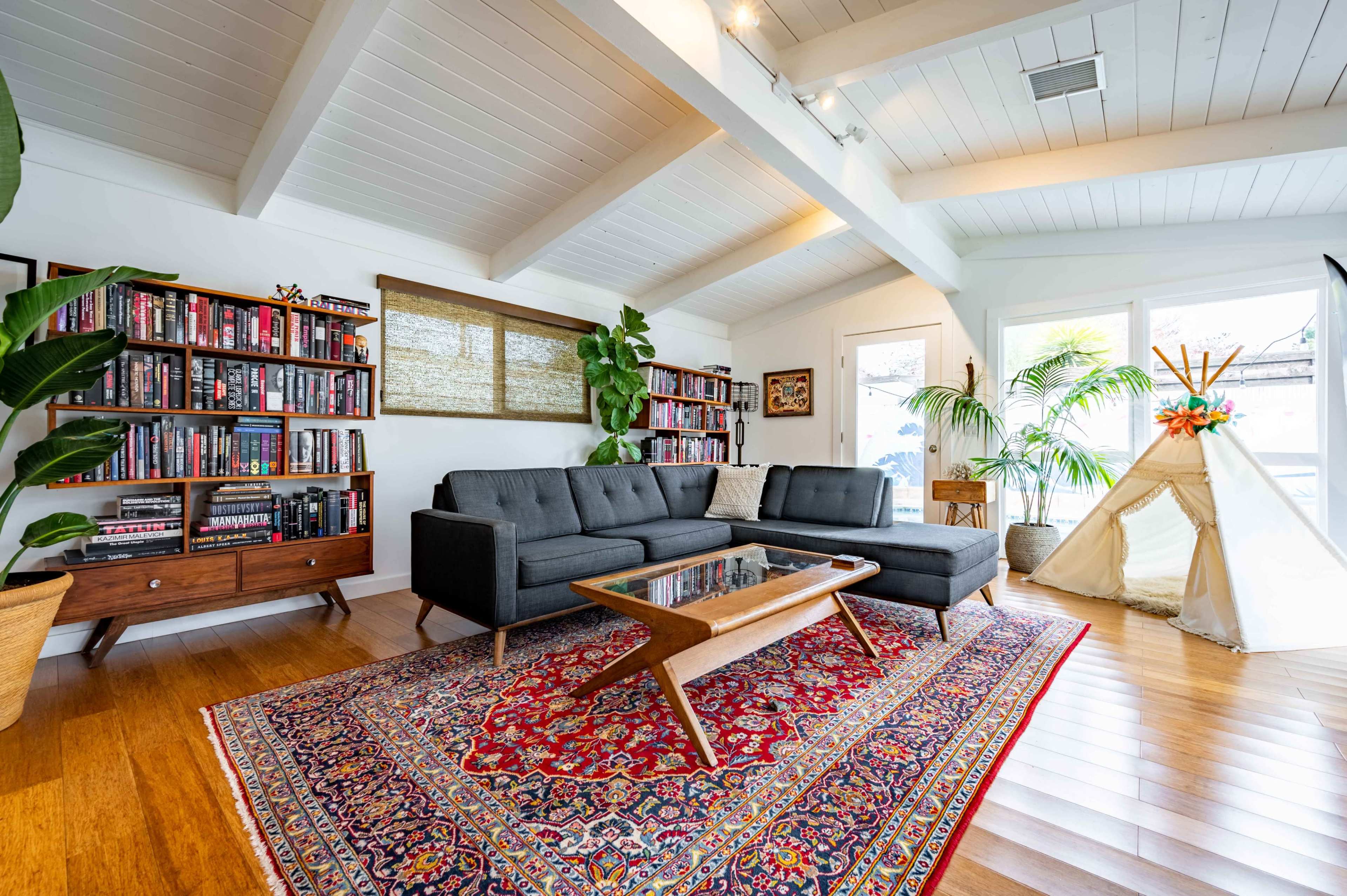 A modern living room with a sectional sofa, a coffee table, bookshelves filled with books, and a children's teepee in the corner.