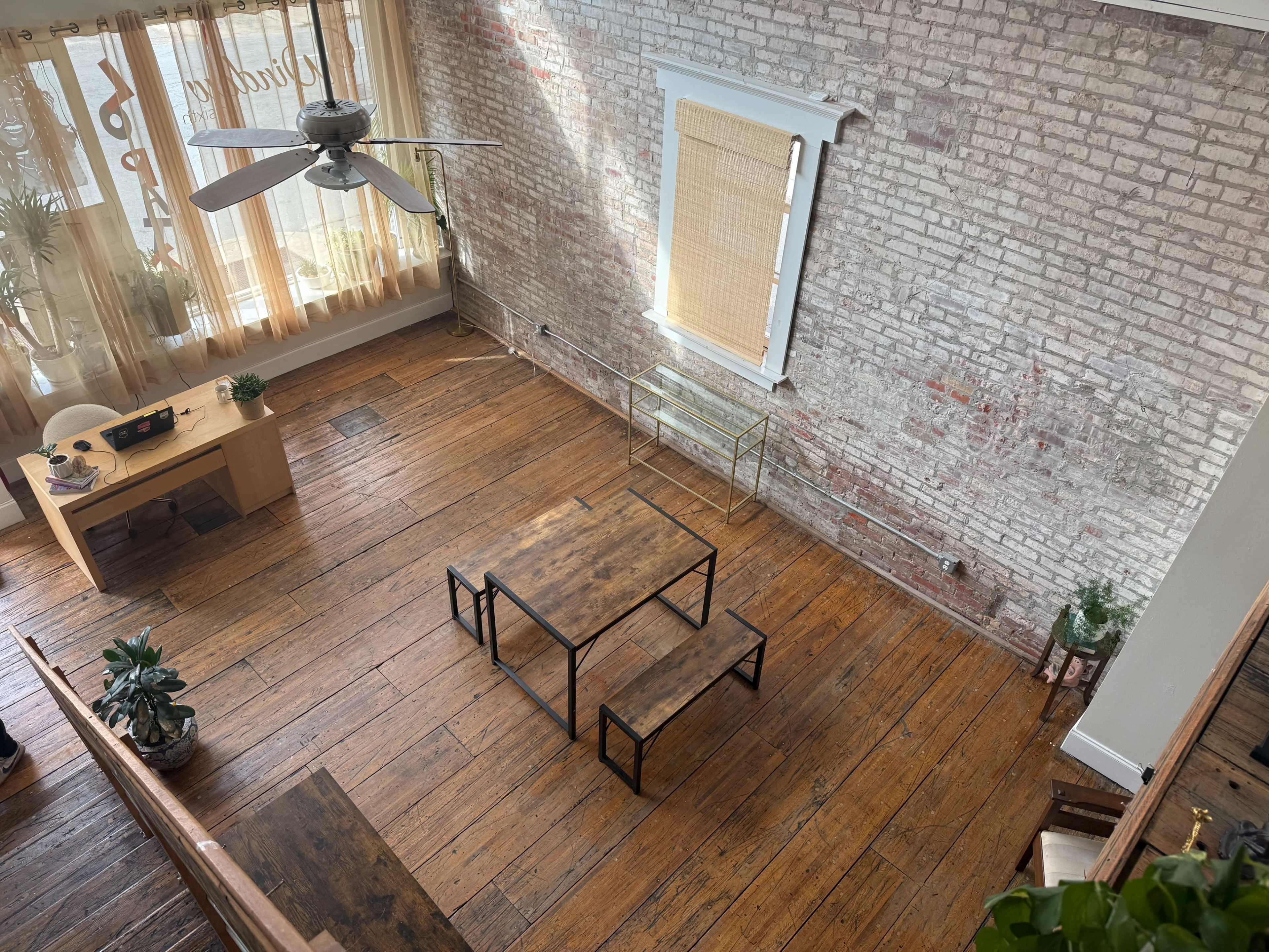 The image shows an indoor space with wooden flooring, a ceiling fan, large windows covered with sheer curtains, and a wooden table accompanied by two black metal benches.