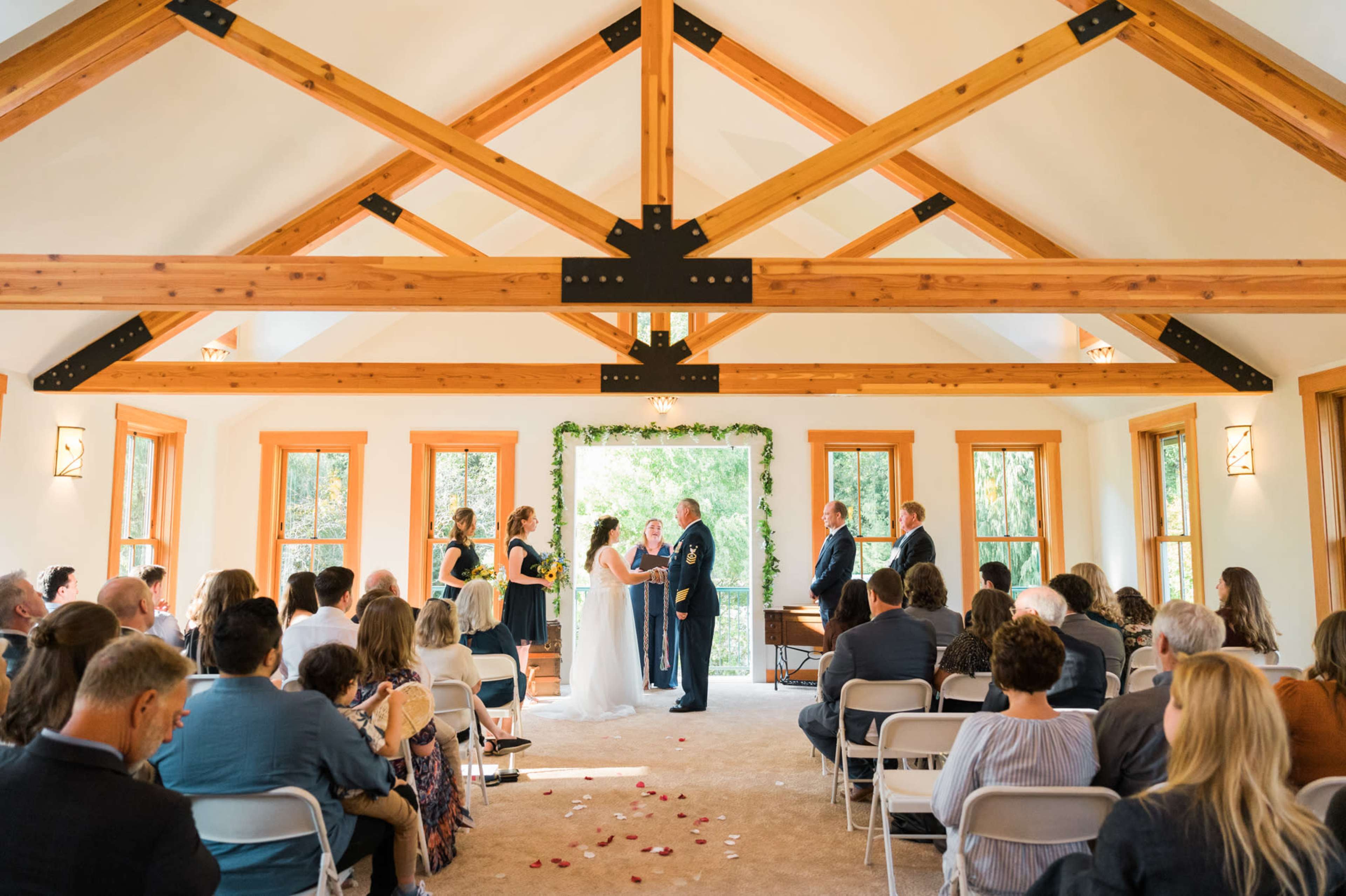 A couple stands at the altar under a floral arch while a wedding ceremony takes place in a well-lit indoor venue surrounded by seated guests.