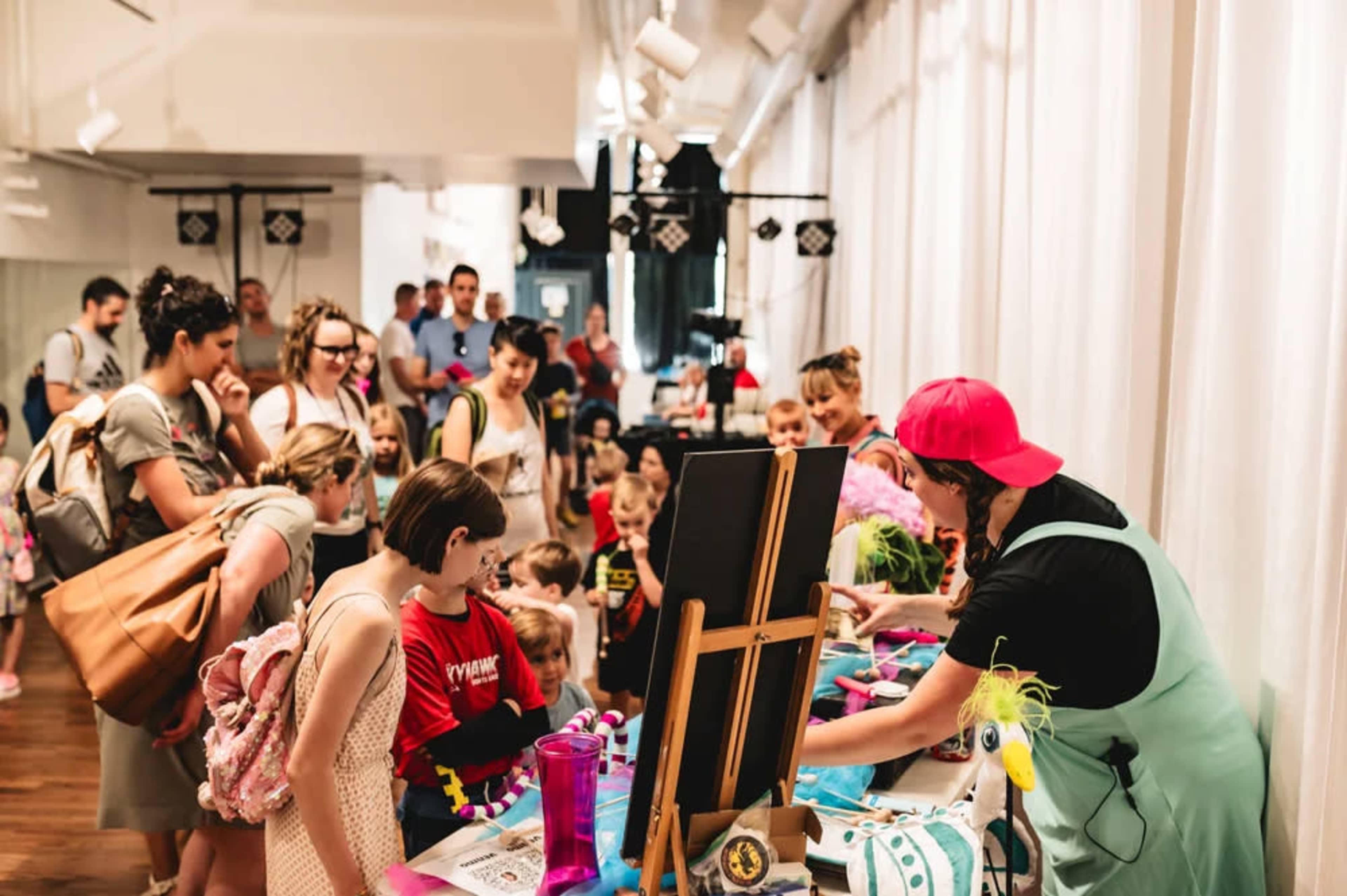A large group of children and adults gathers around a table where an individual in a colorful outfit is displaying art supplies and activities.