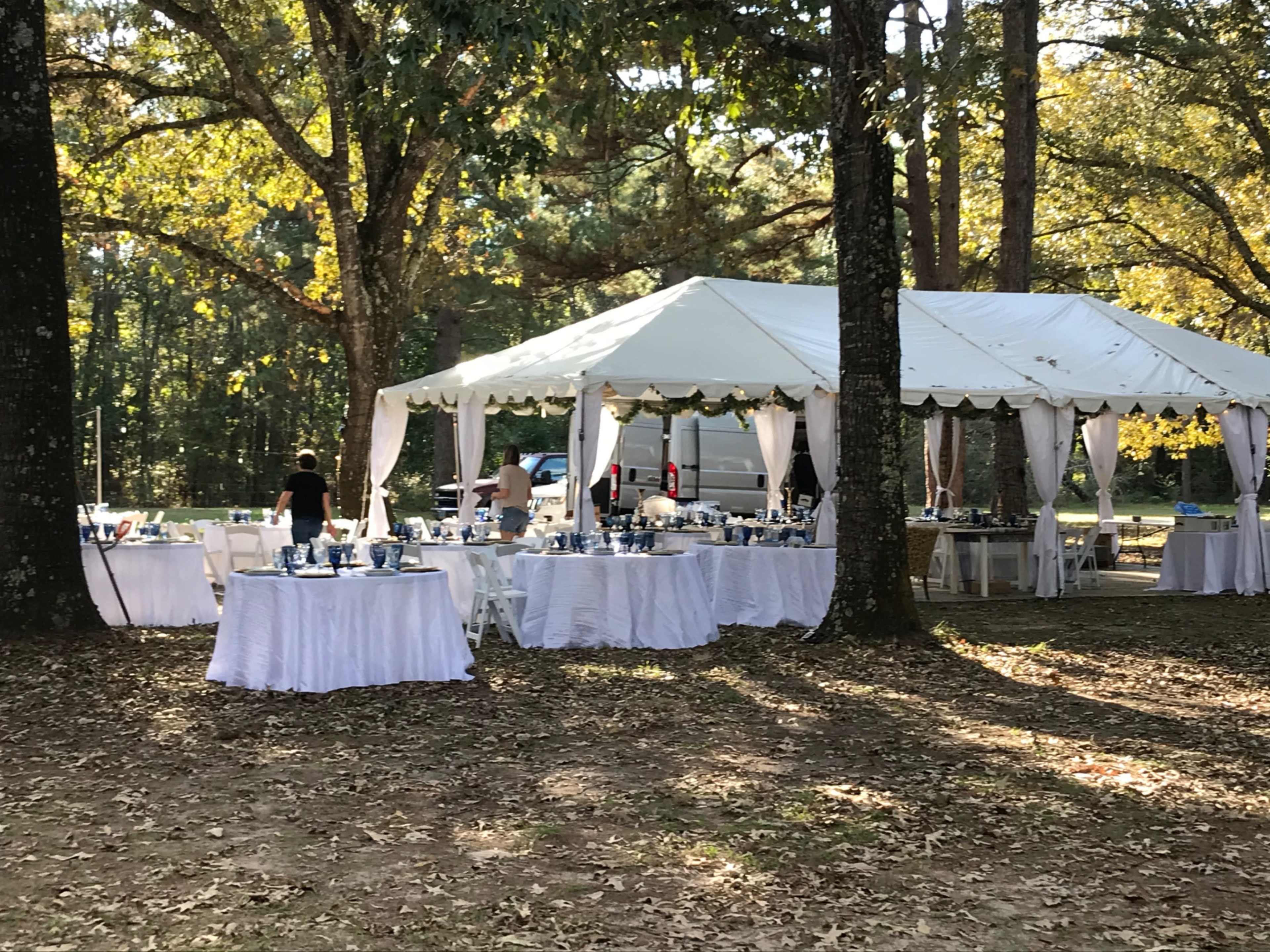 A large white tent is set up in a wooded area, surrounded by tables covered with white tablecloths and various diningware.