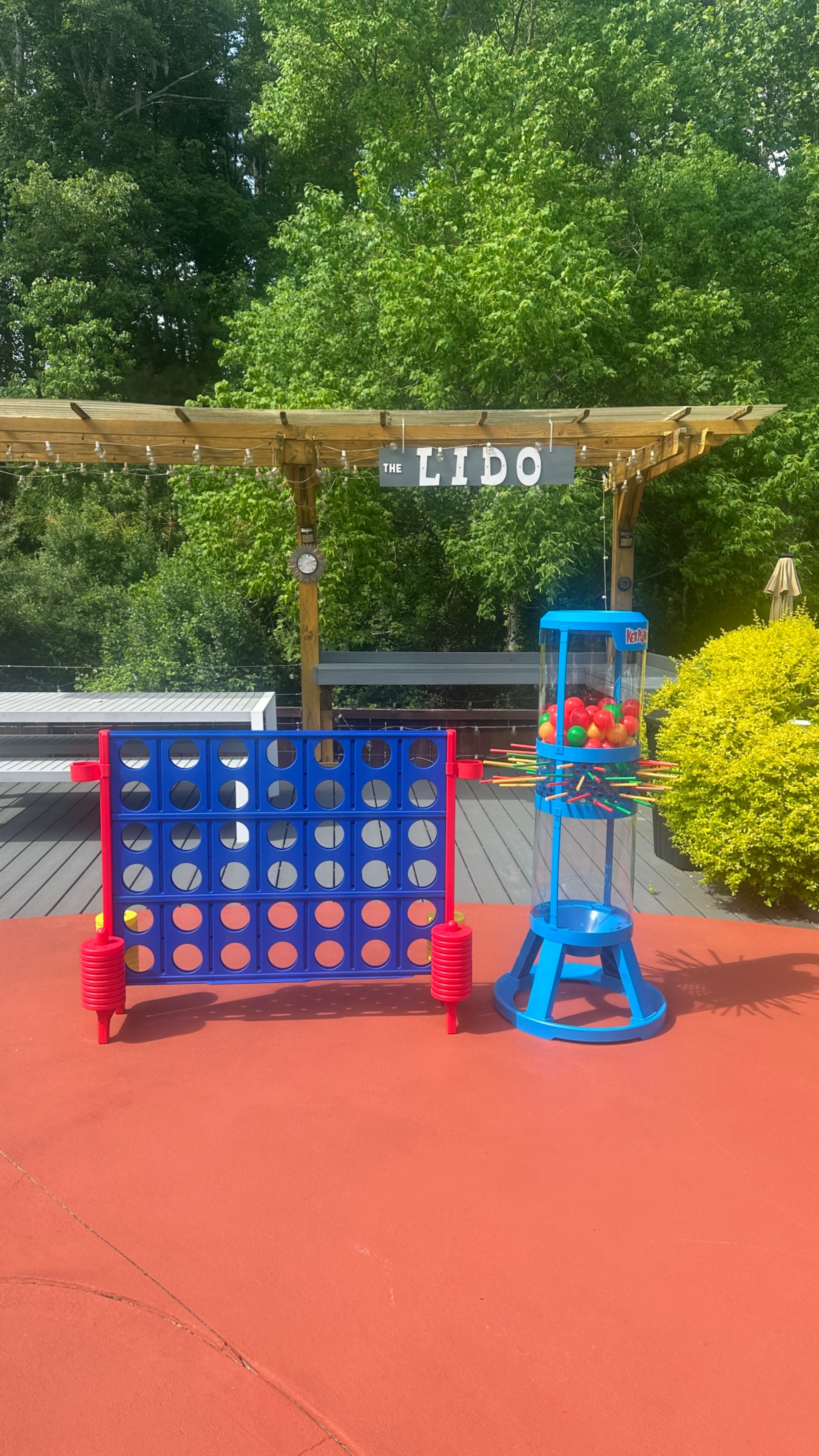 The image shows a colorful outdoor setup featuring a large Connect Four game on the left and a gumball machine on the right, with a wooden pergola labeled "The Lido" in the background.