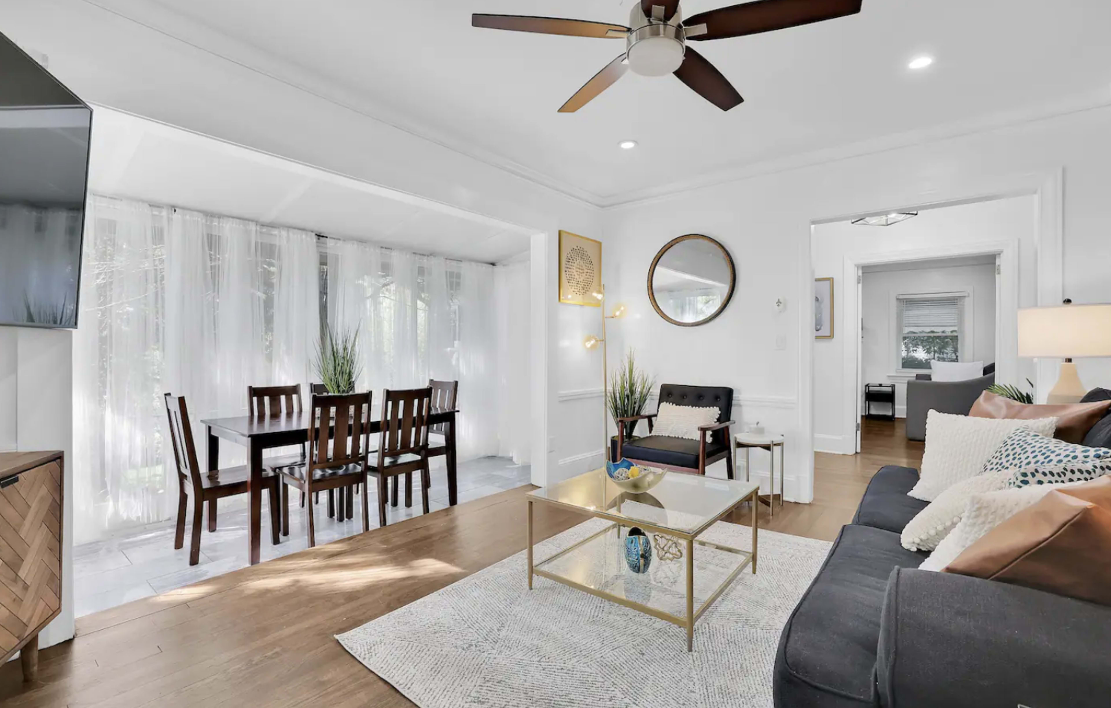 A bright, modern living room with a seating area, a dining table nearby, and large windows with sheer curtains allowing natural light to enter.