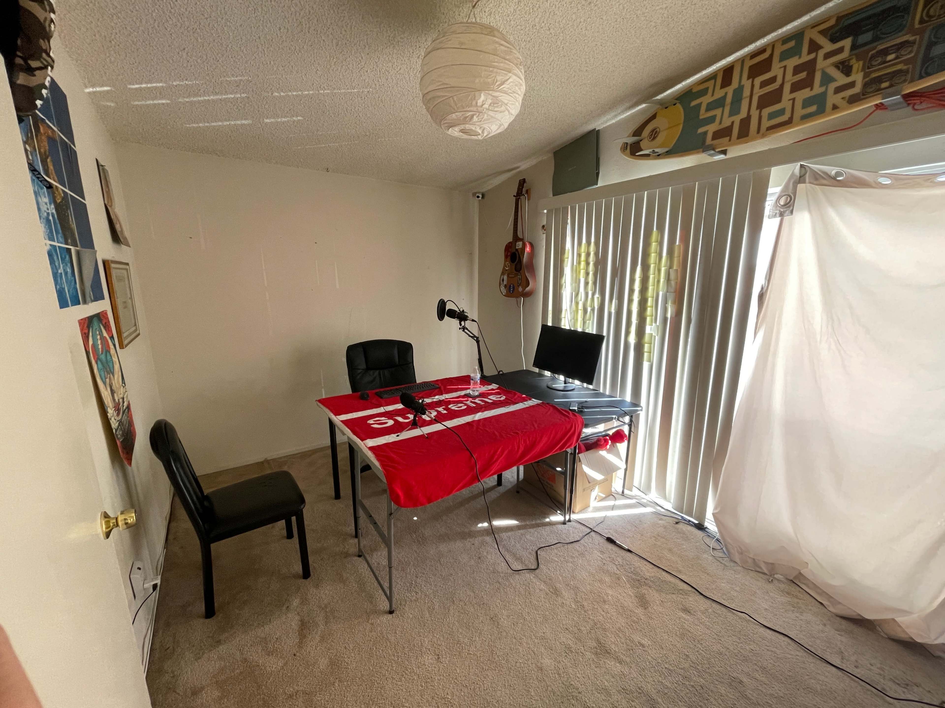 A small room with a table covered by a red blanket, two black chairs, a microphone set up, and blinds partially open to let in light.