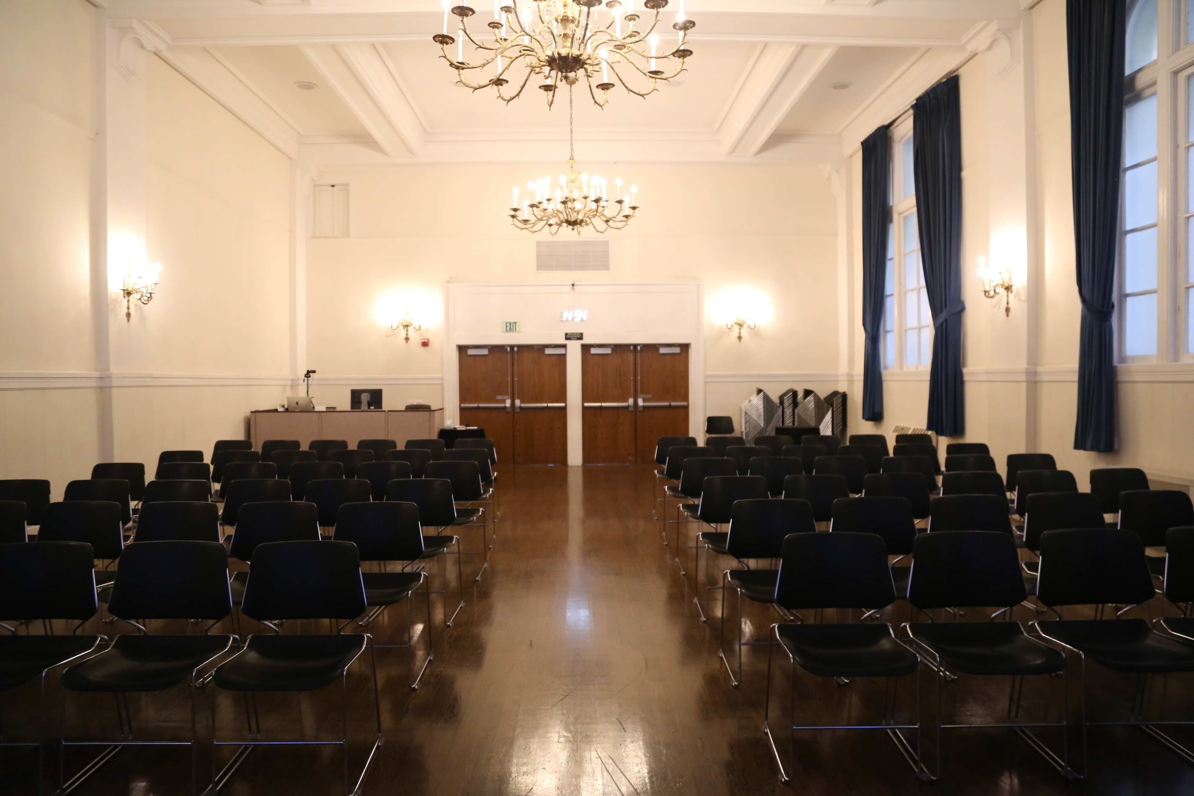 An empty auditorium with rows of black chairs facing a front stage area, featuring a chandelier and large windows.