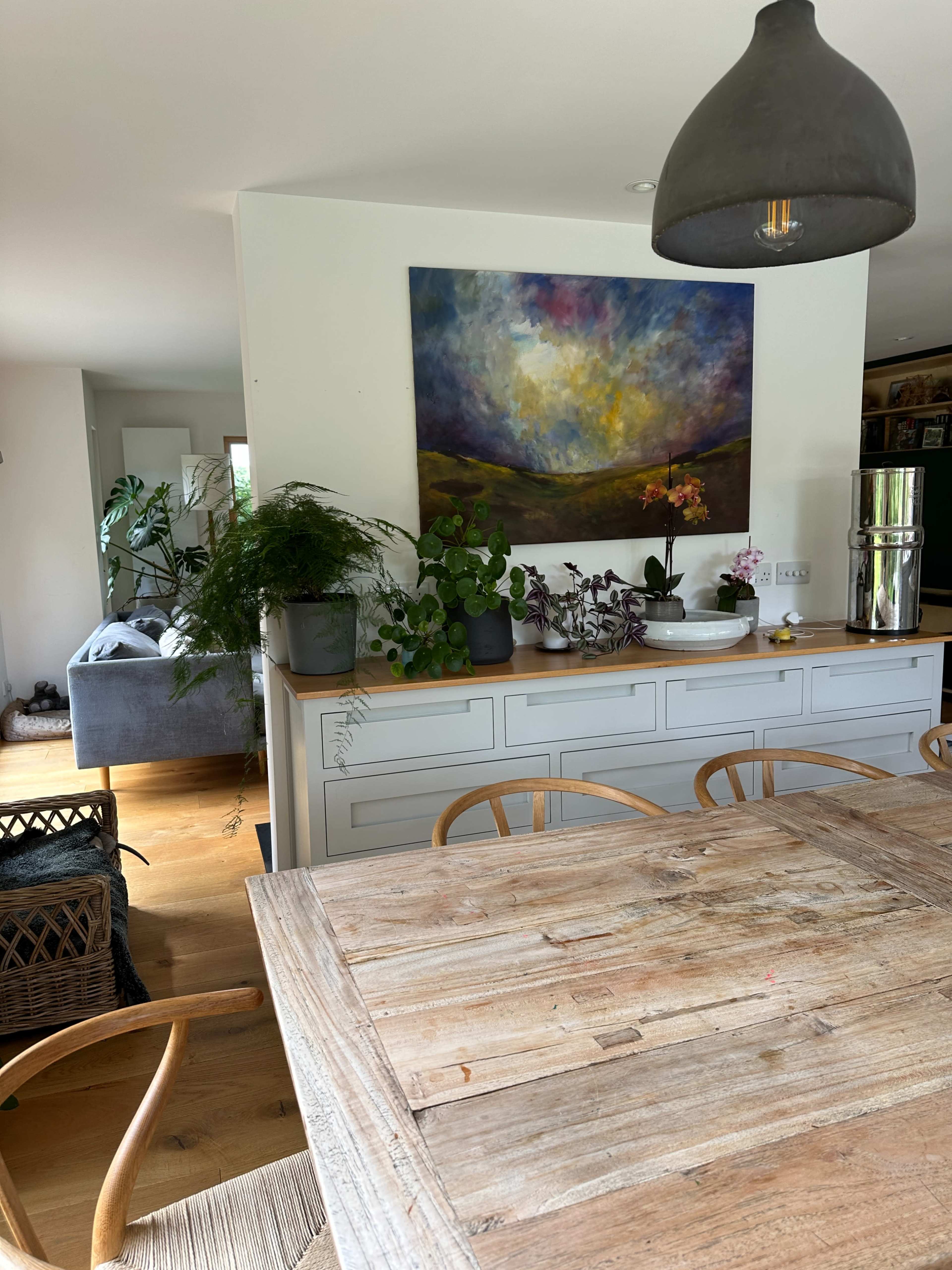 A dining area features a wooden table surrounded by chairs, with a large painting and various plants displayed on a white sideboard.