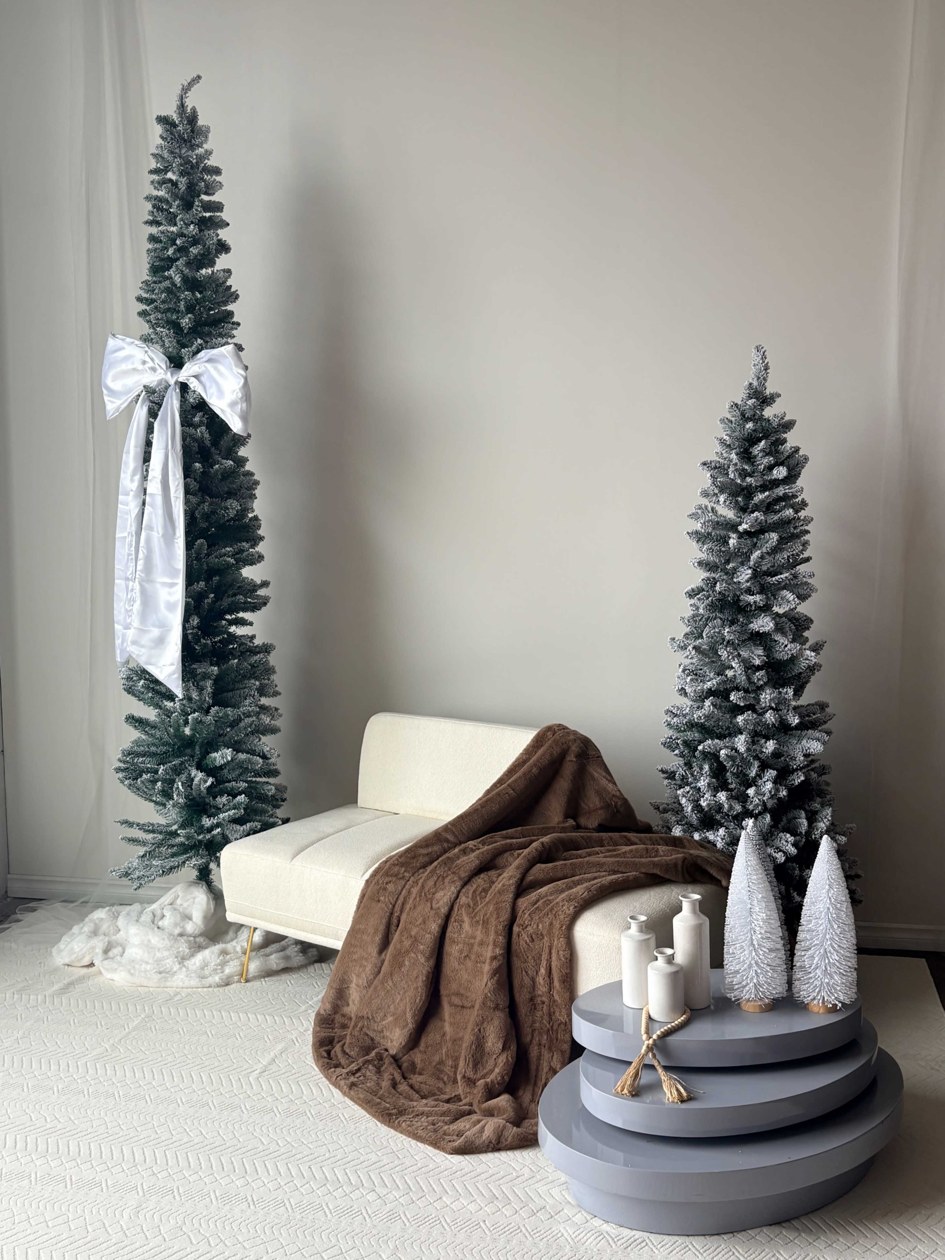 The image features two decorated Christmas trees, a white couch, a brown throw blanket, and three candles arranged on a tiered gray table.